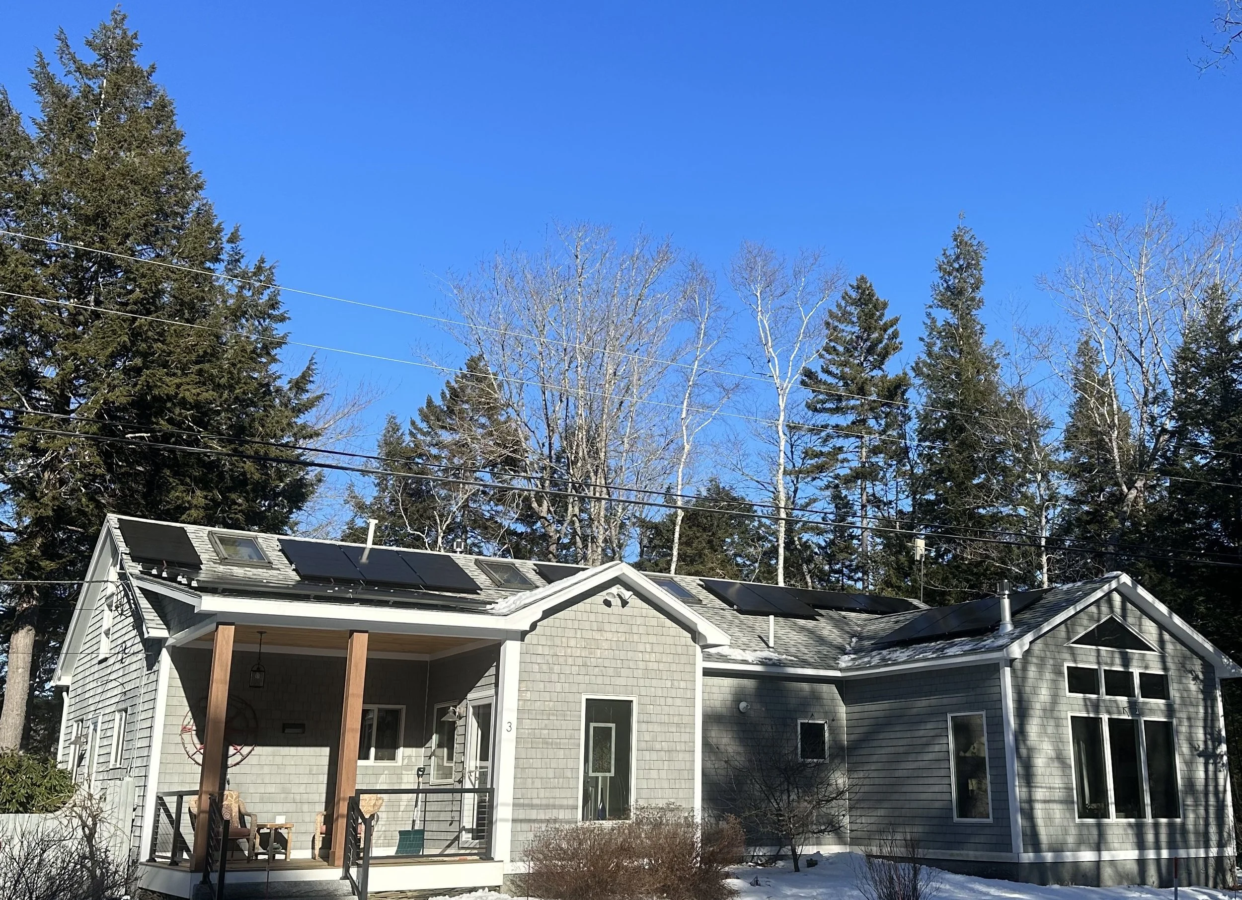 A two-story house with solar panels on the roof, gray siding, large windows, a front porch with wooden posts, and a patio with outdoor furniture. There are trees and a clear blue sky in the background, with some snow on the ground.