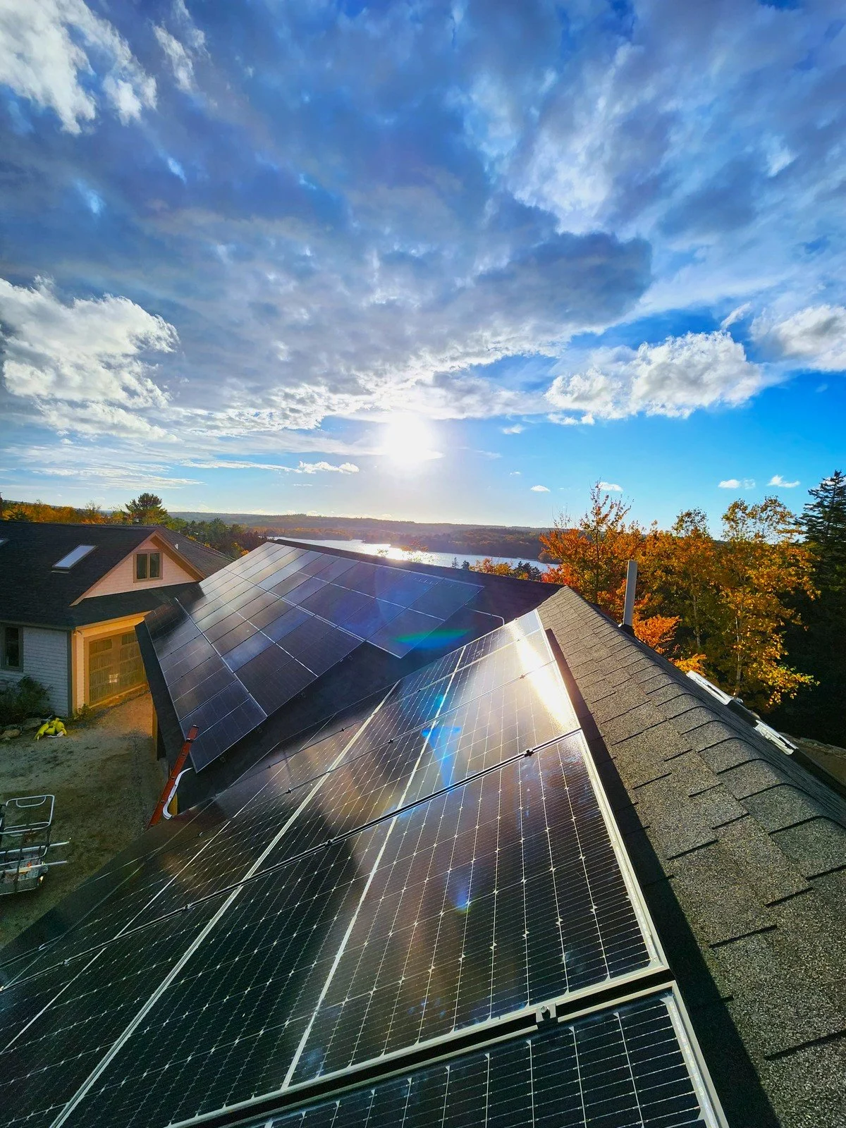 House roof with solar panels during sunset, trees with autumn foliage, lake and sky with clouds in the background.