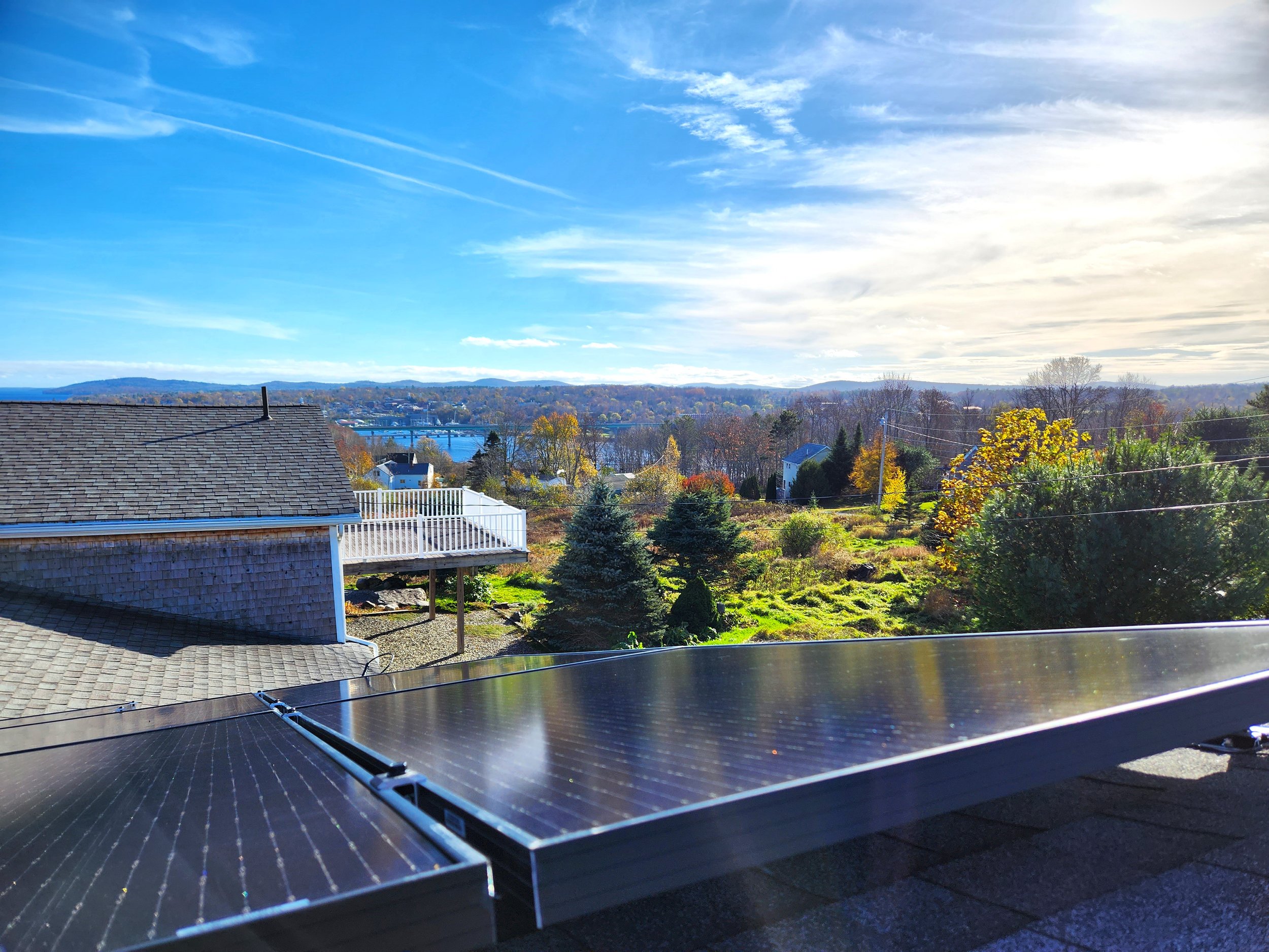 A scenic view of a neighborhood with rooftops, trees showing fall colors, a river in the middle distance, and a partly cloudy sky.
