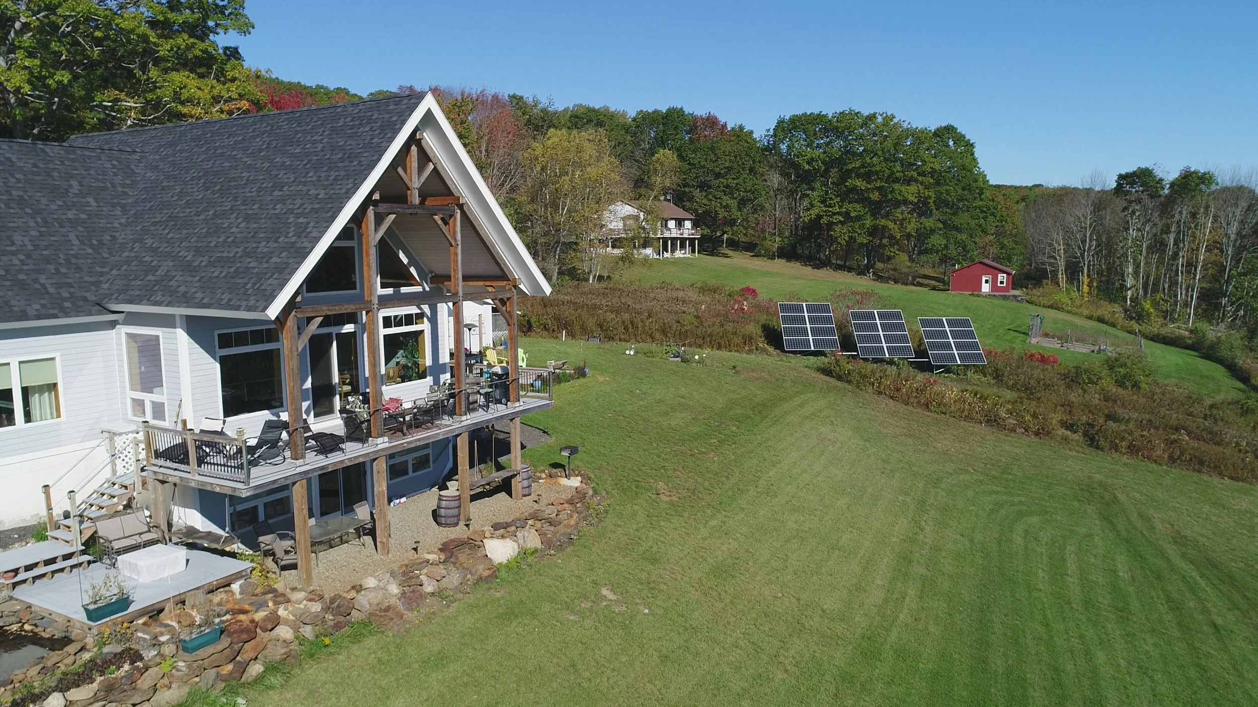 A white house with a dark gray gabled roof, large wooden supports, and a wraparound deck with outdoor furniture, surrounded by green grass and trees, with solar panels on a hillside and a small red shed in the distance under a clear blue sky.