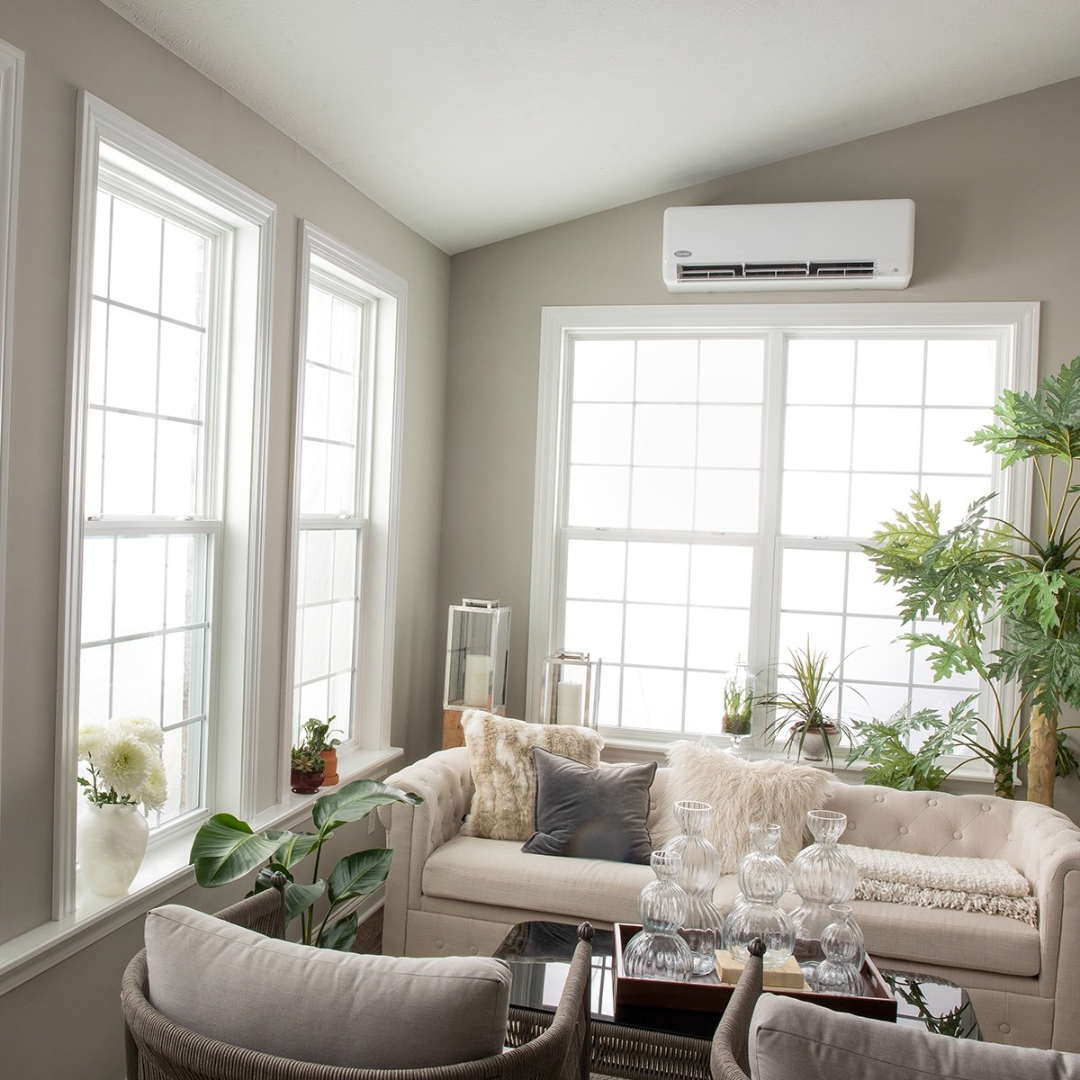 Bright living room with beige sofas, glass vases on a tray, and multiple large windows with white trim, decorated with potted plants and candles.