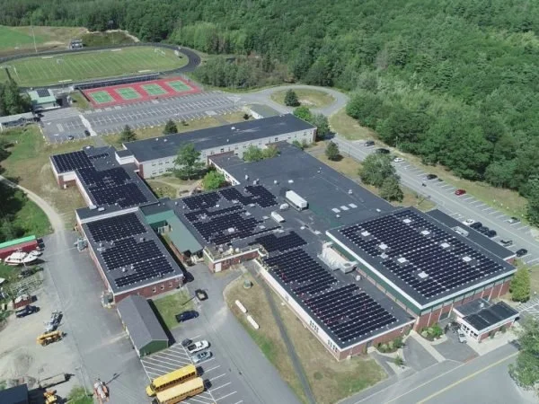 An aerial view of a school campus with solar panels on the rooftops, parking lot, and sports fields surrounded by trees.