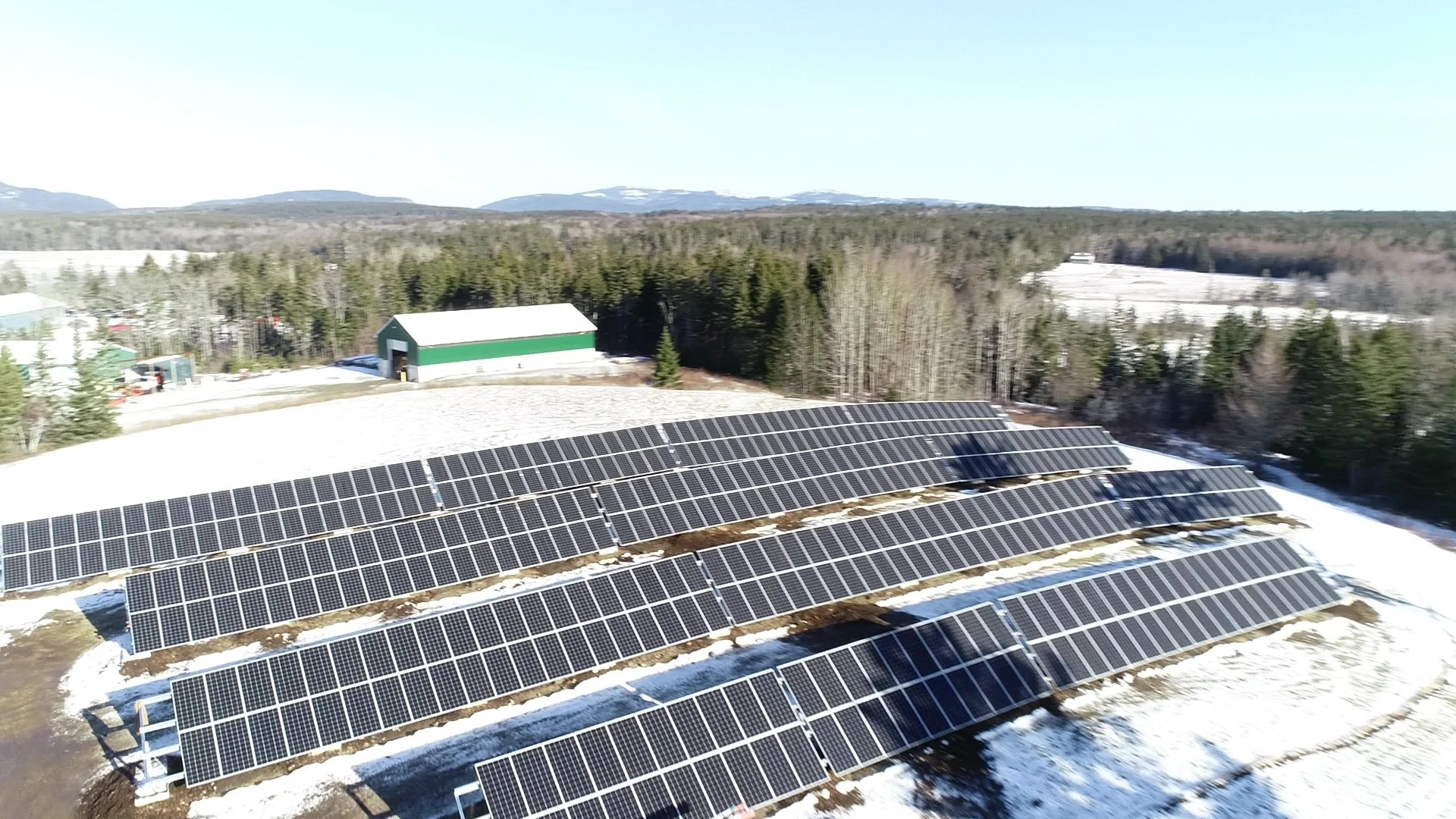 Aerial view of a solar panel array in a snowy landscape with trees, mountains, and farm buildings in the background.