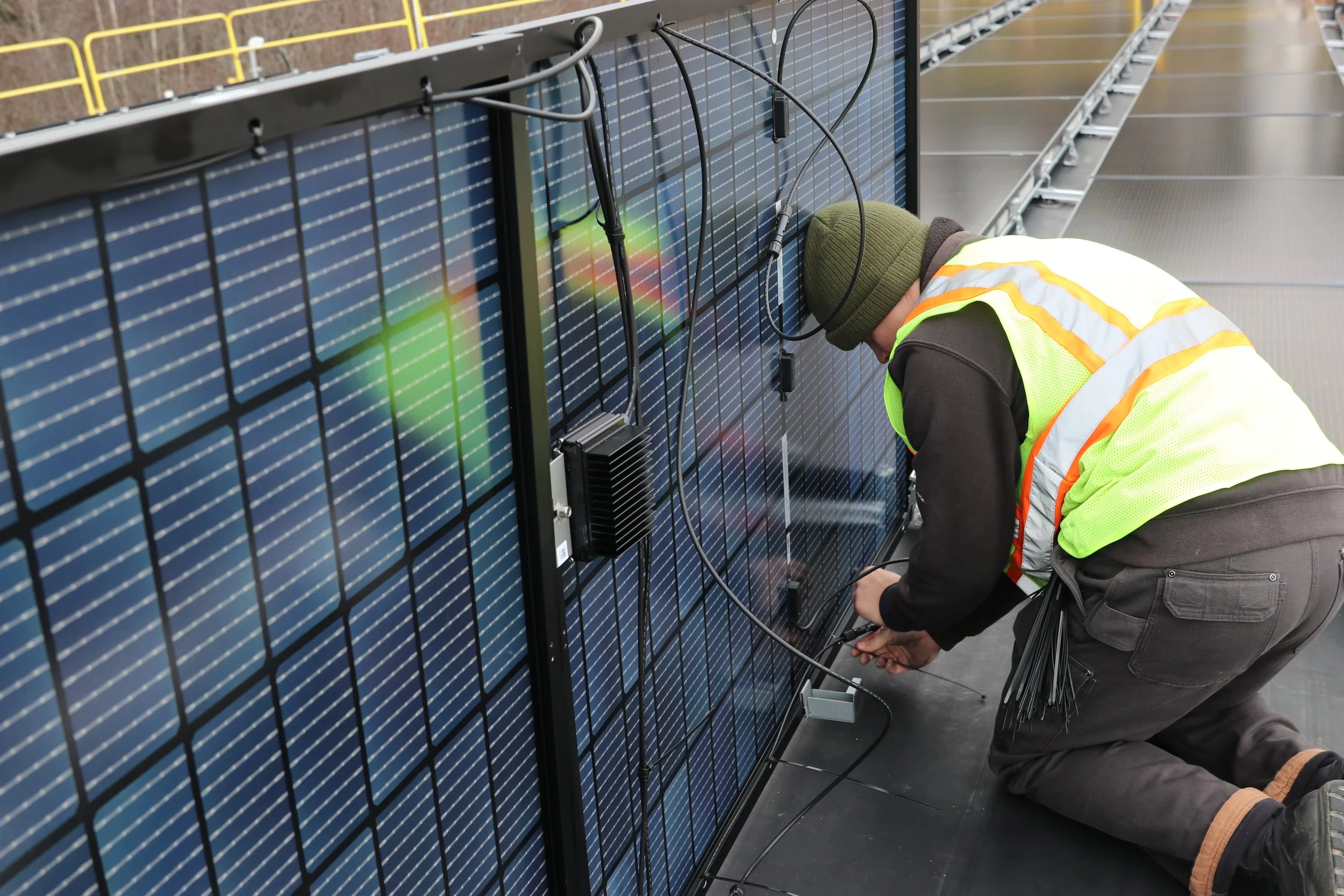 A person in a yellow safety vest and olive green beanie working on a large solar panel, kneeling on a dark floor with tools connected to the panel.