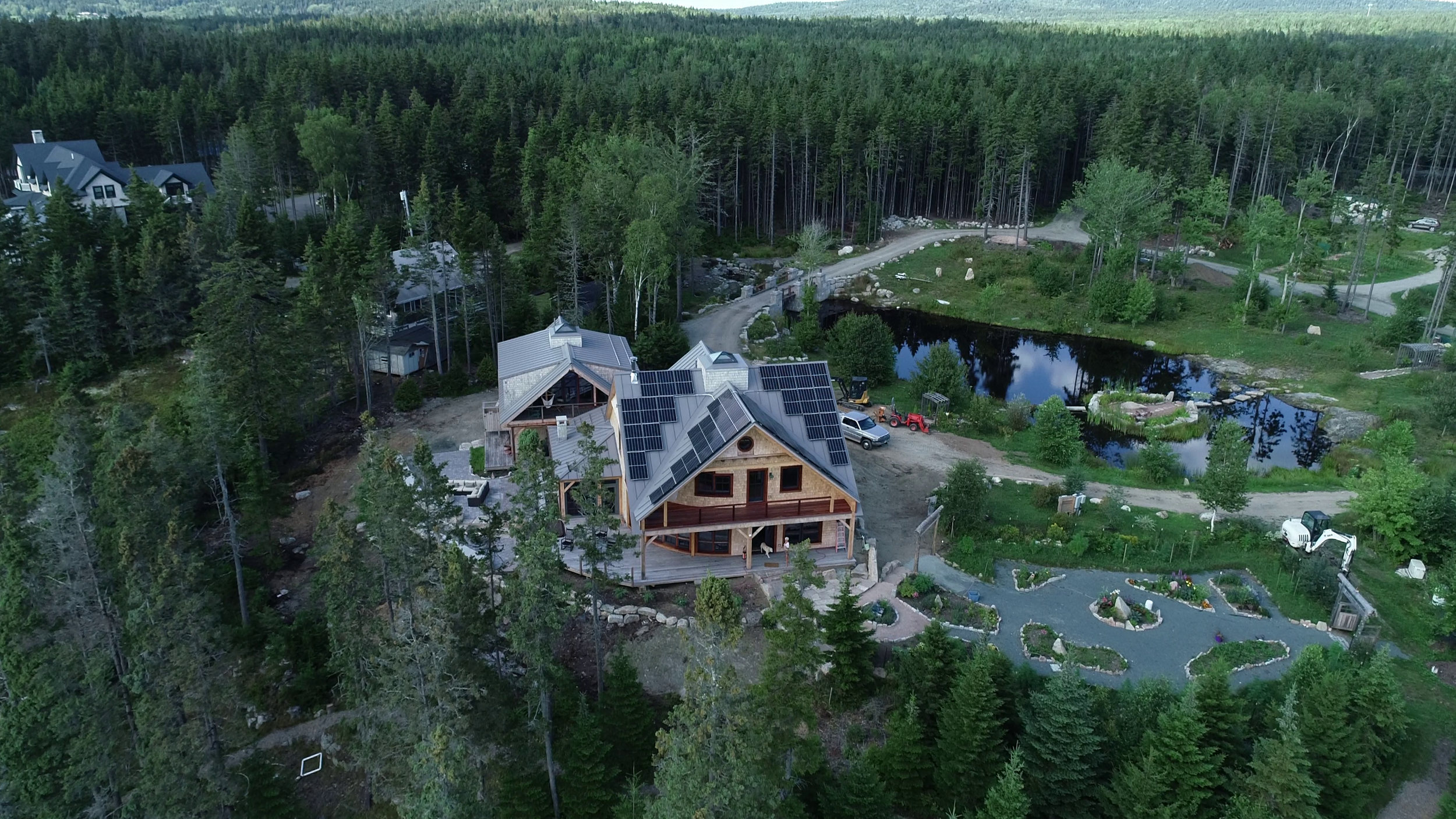 Aerial view of a residential house under construction in a scenic forested area, featuring solar panels on the roof, a pond nearby, and a winding driveway with construction vehicles.