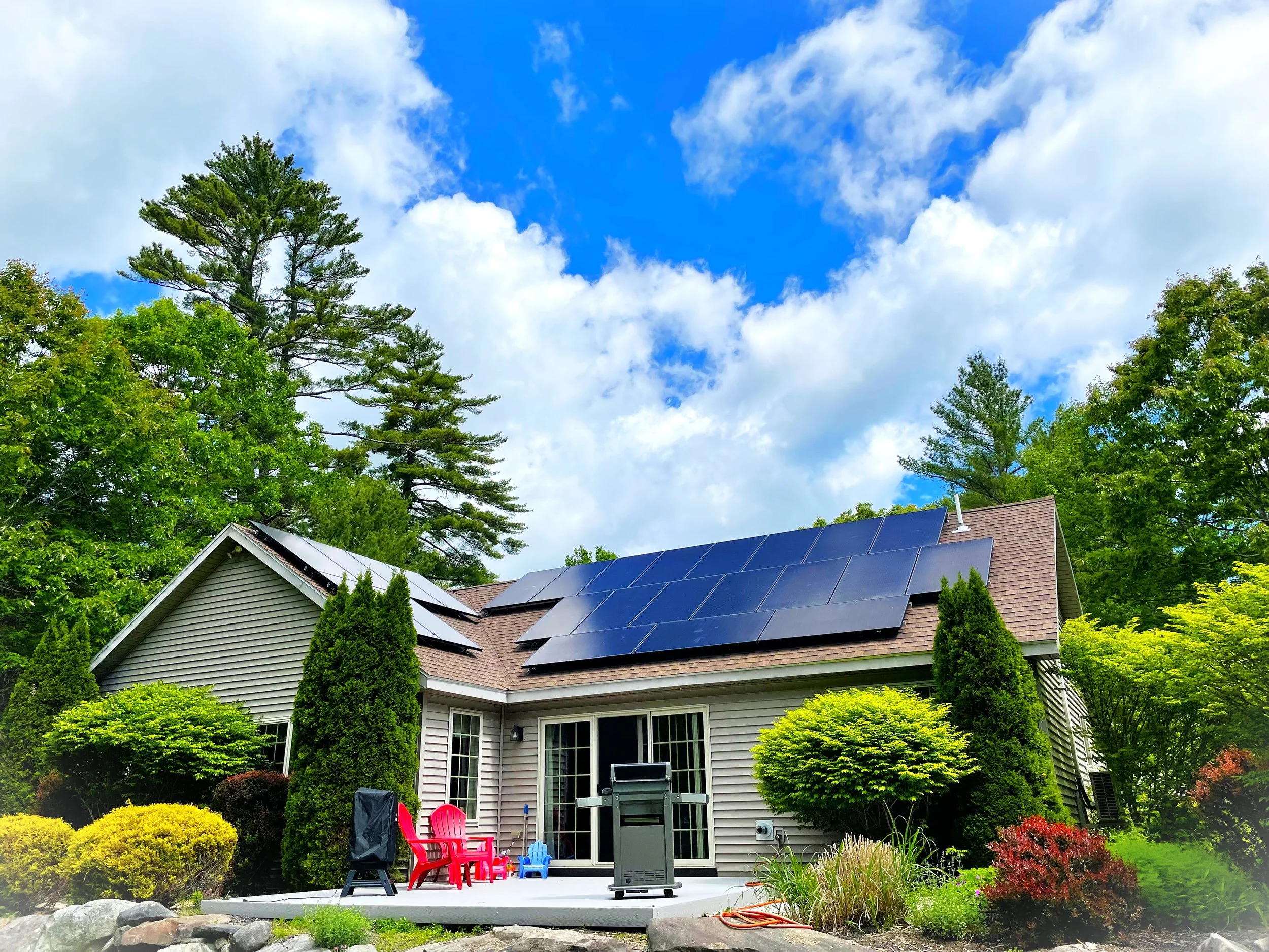 Backyard patio with patio furniture, grill, and garden, house with solar panels on roof, trees, and a partly cloudy sky