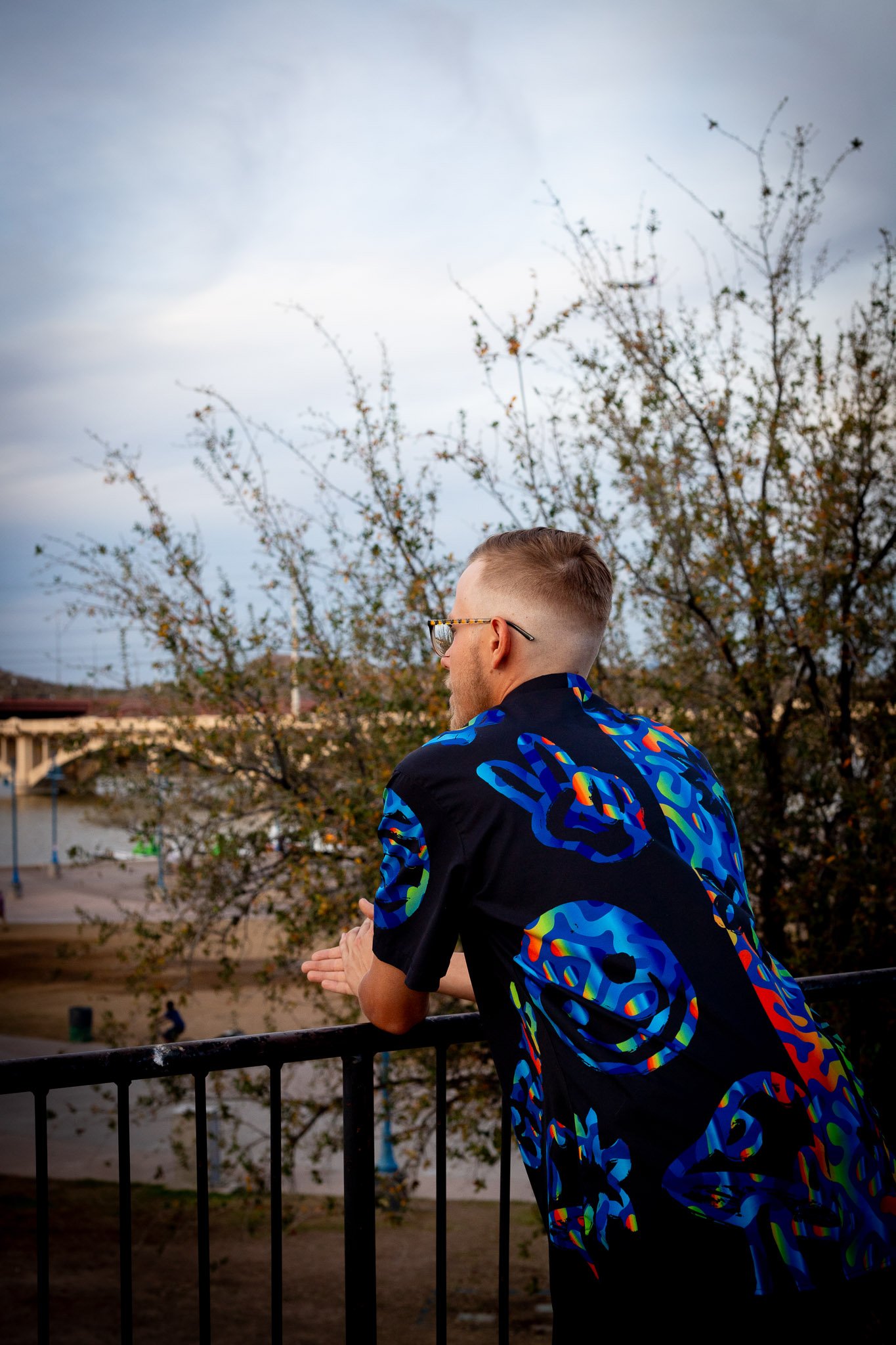 A man with short light hair and glasses leans on a black railing, looking out at a park with trees and a bridge in the background, wearing a black shirt with colorful psychedelic designs.