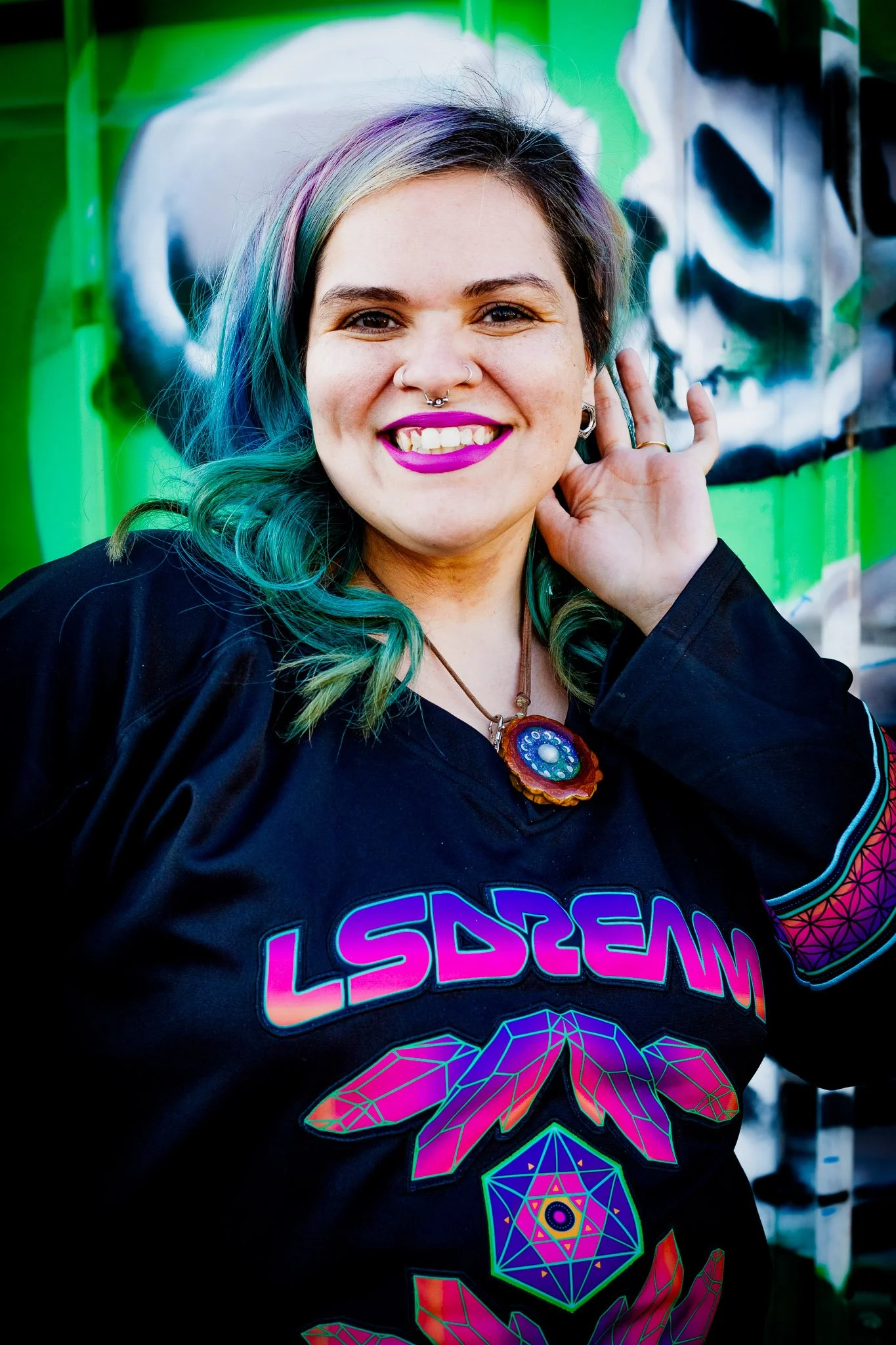A woman with rainbow-colored hair, bright pink lipstick, and multiple facial piercings, smiling and touching her ear, standing in front of a graffiti wall.