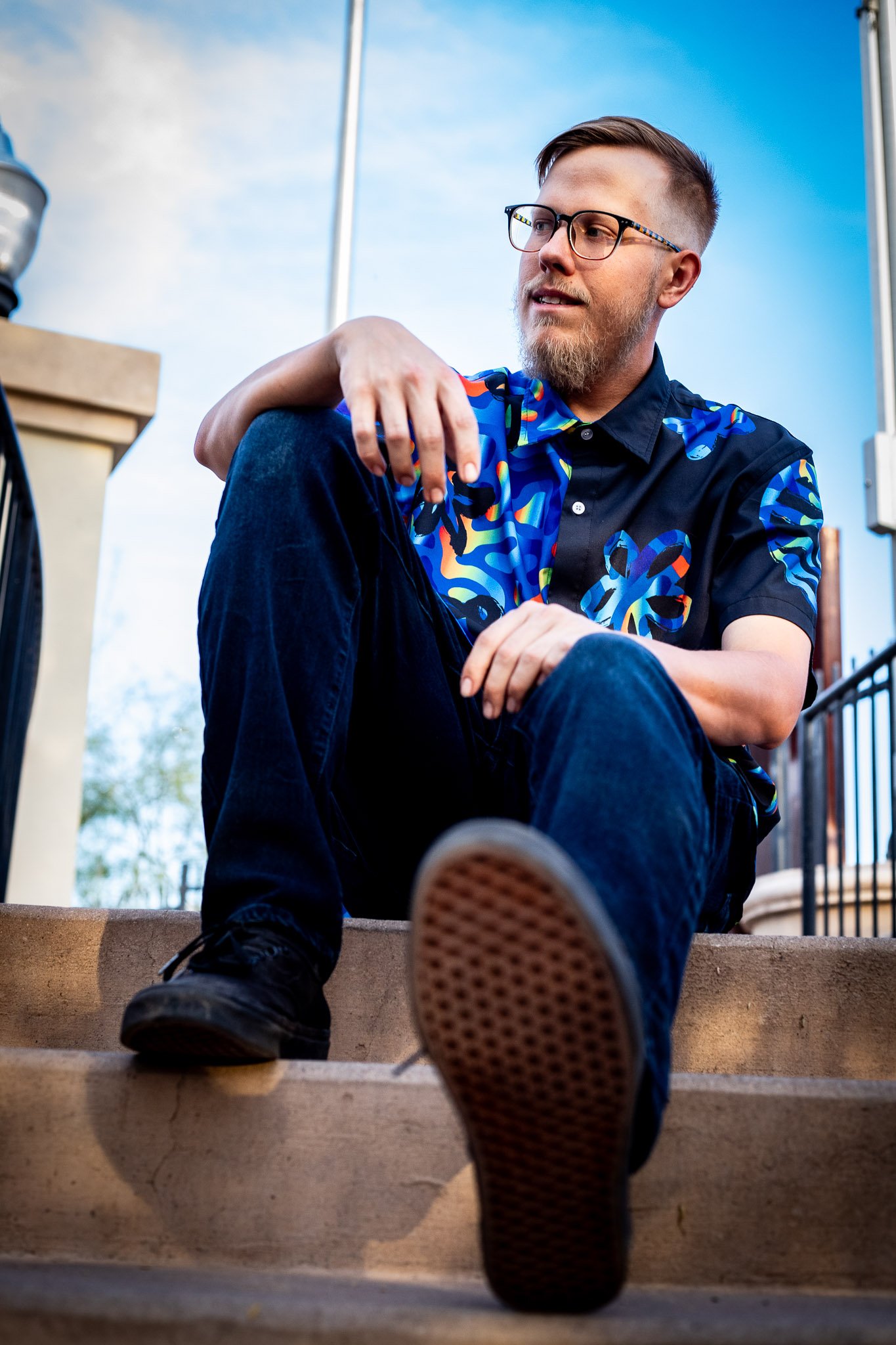 A man with glasses and a beard sitting on outdoor stairs, looking to his left, wearing a blue patterned short-sleeve shirt, black pants, and black shoes.