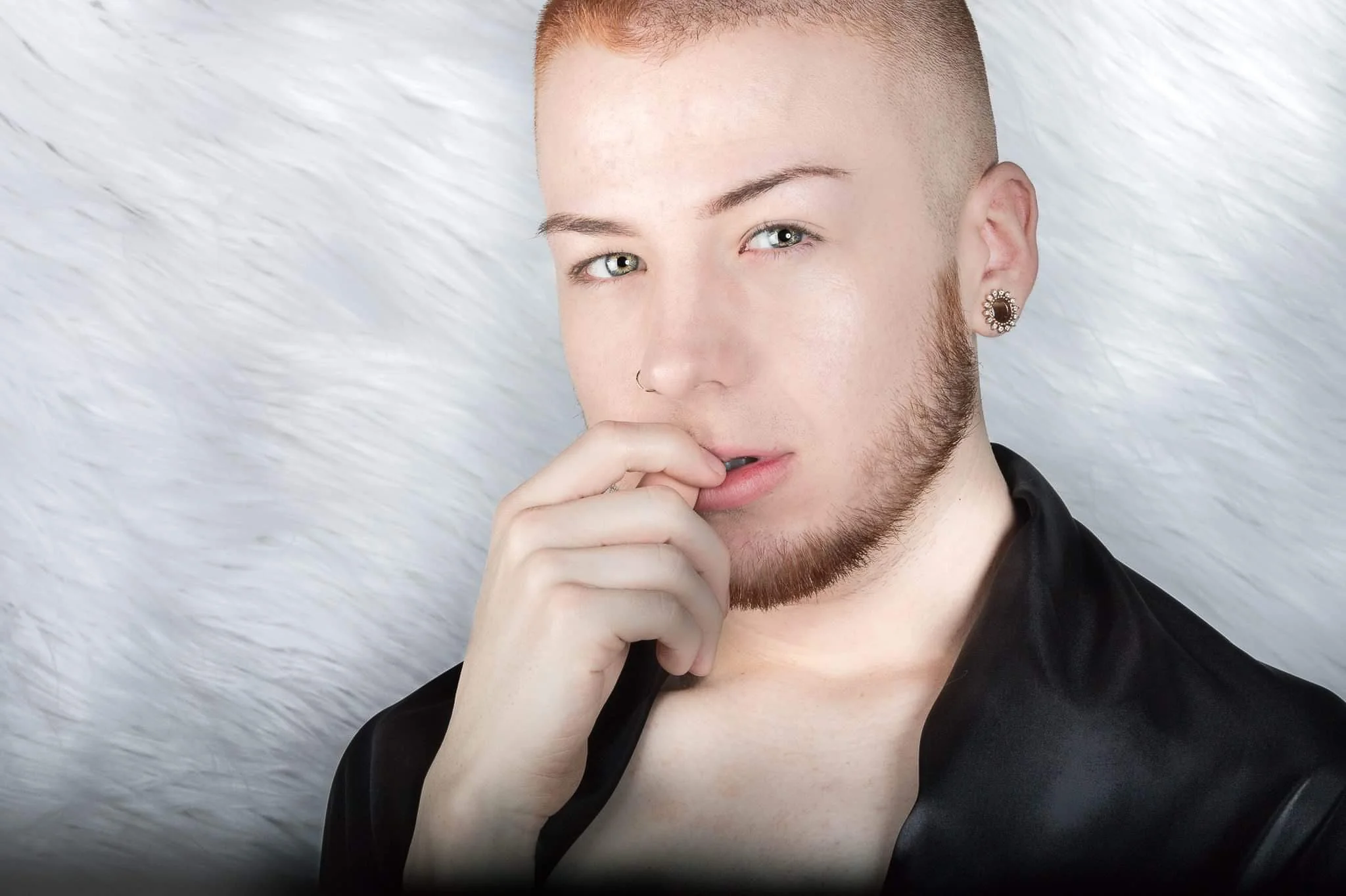 A young person with short hair and a beard, looking at the camera, biting their finger, with a soft white background.