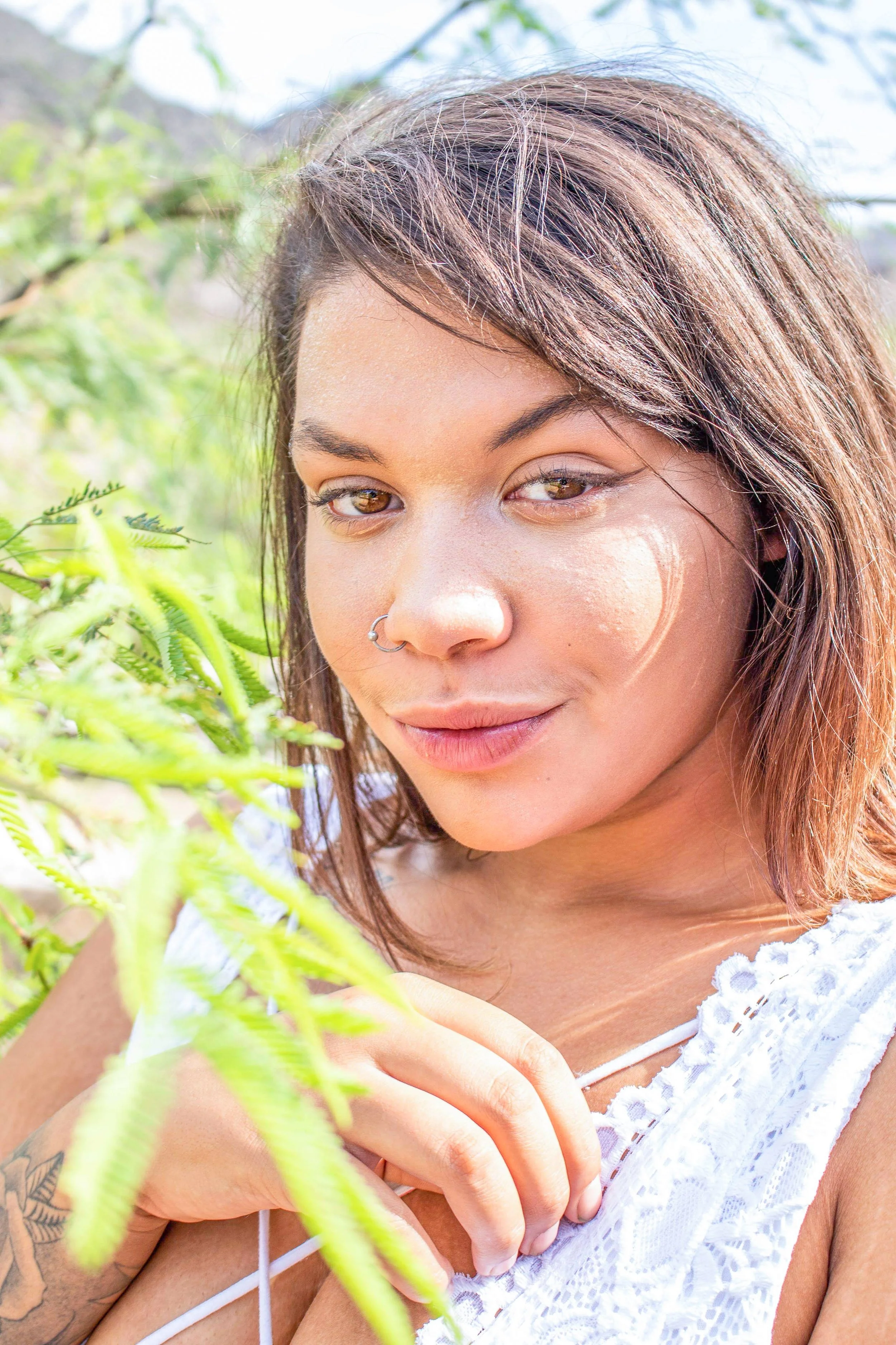 A young woman with brown hair, wearing a white lace top, posing outdoors near greenery and sunlight.