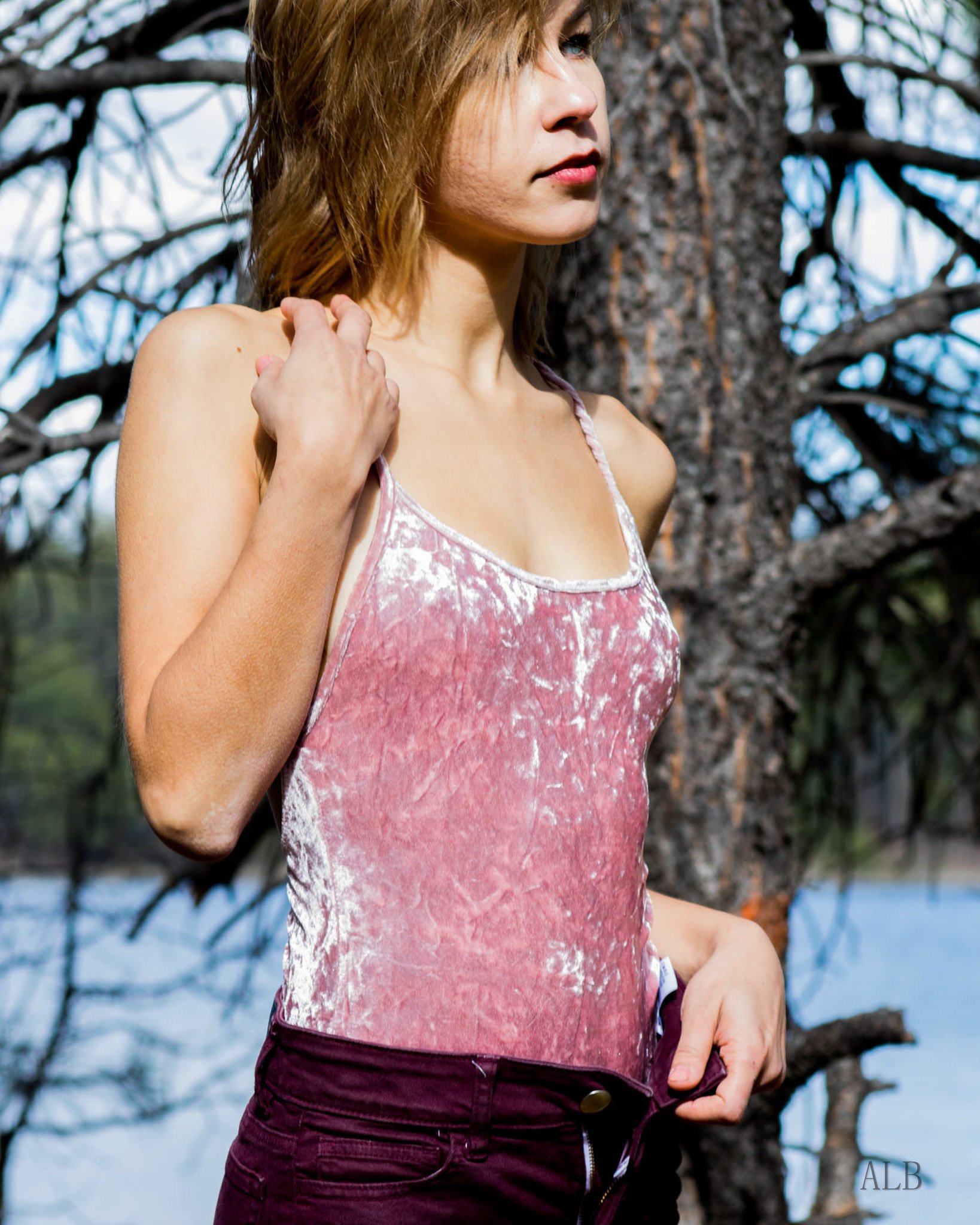 A woman with blonde hair wearing a pink velvet top standing outdoors near a tree with branches, with water in the background.