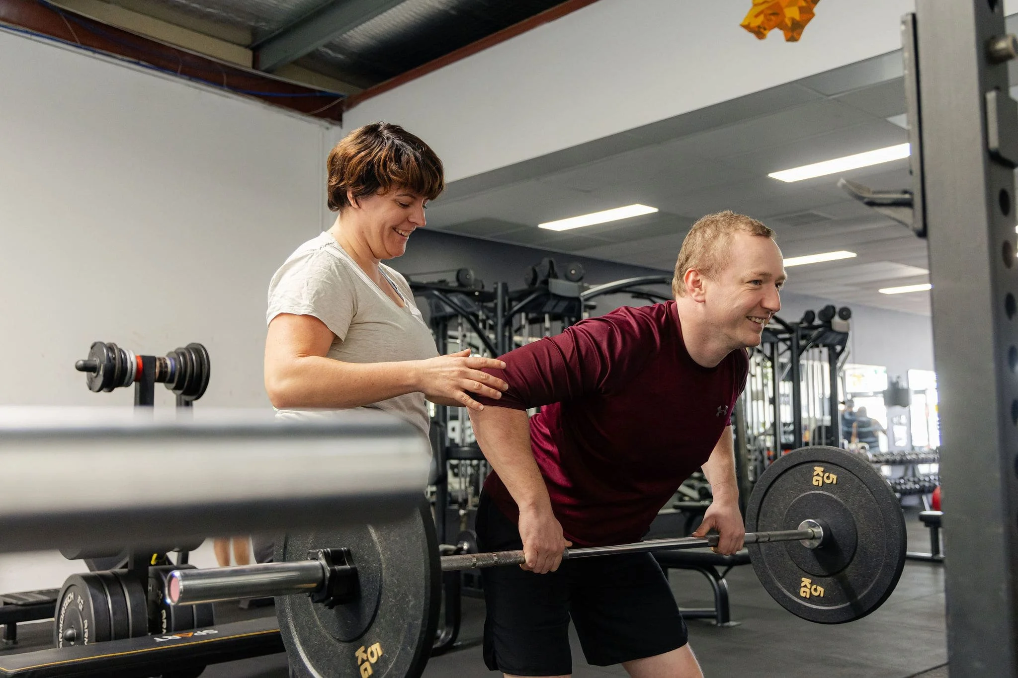 Adventure Physio, Stephanie Lock assisting a man lifting a barbell during a workout in a gym during an injury rehabilitation session at Transpire Fitness in Bibra Lake