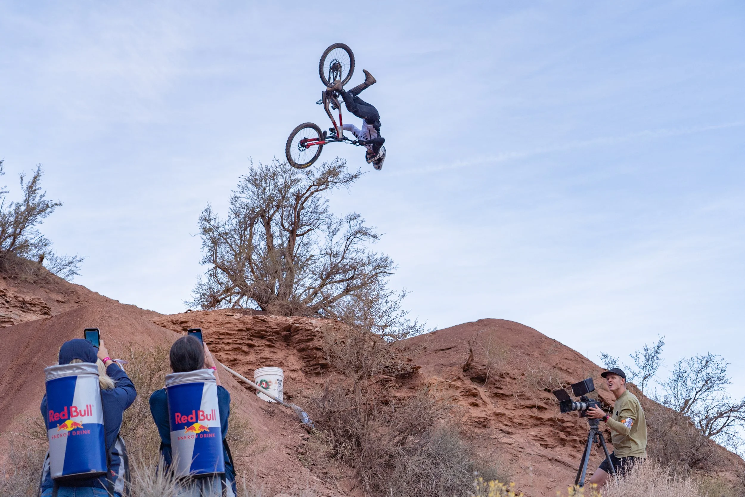 Mountain biker performing a flip off a dirt jump while being photographed by people with Red Bull gear.