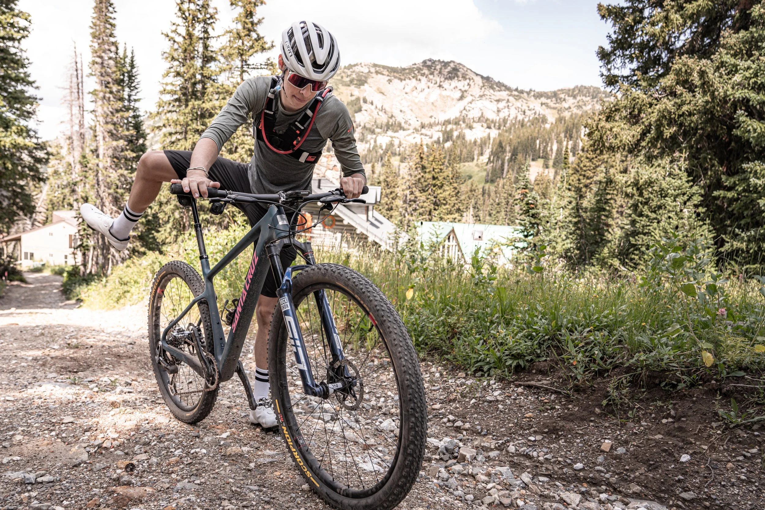 Person with a helmet and sunglasses mounting a mountain bike on a rocky trail surrounded by trees and mountains. | Photography by Anders Henrikson Photo 