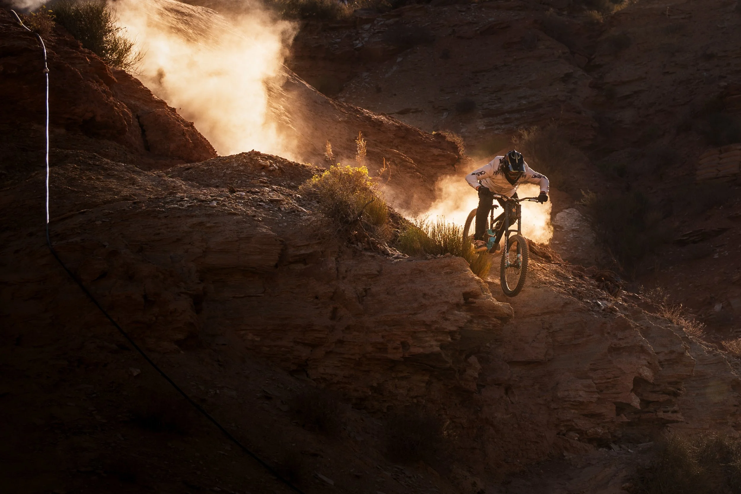 Mountain biker riding down rocky terrain with dust clouds in the background during sunset.