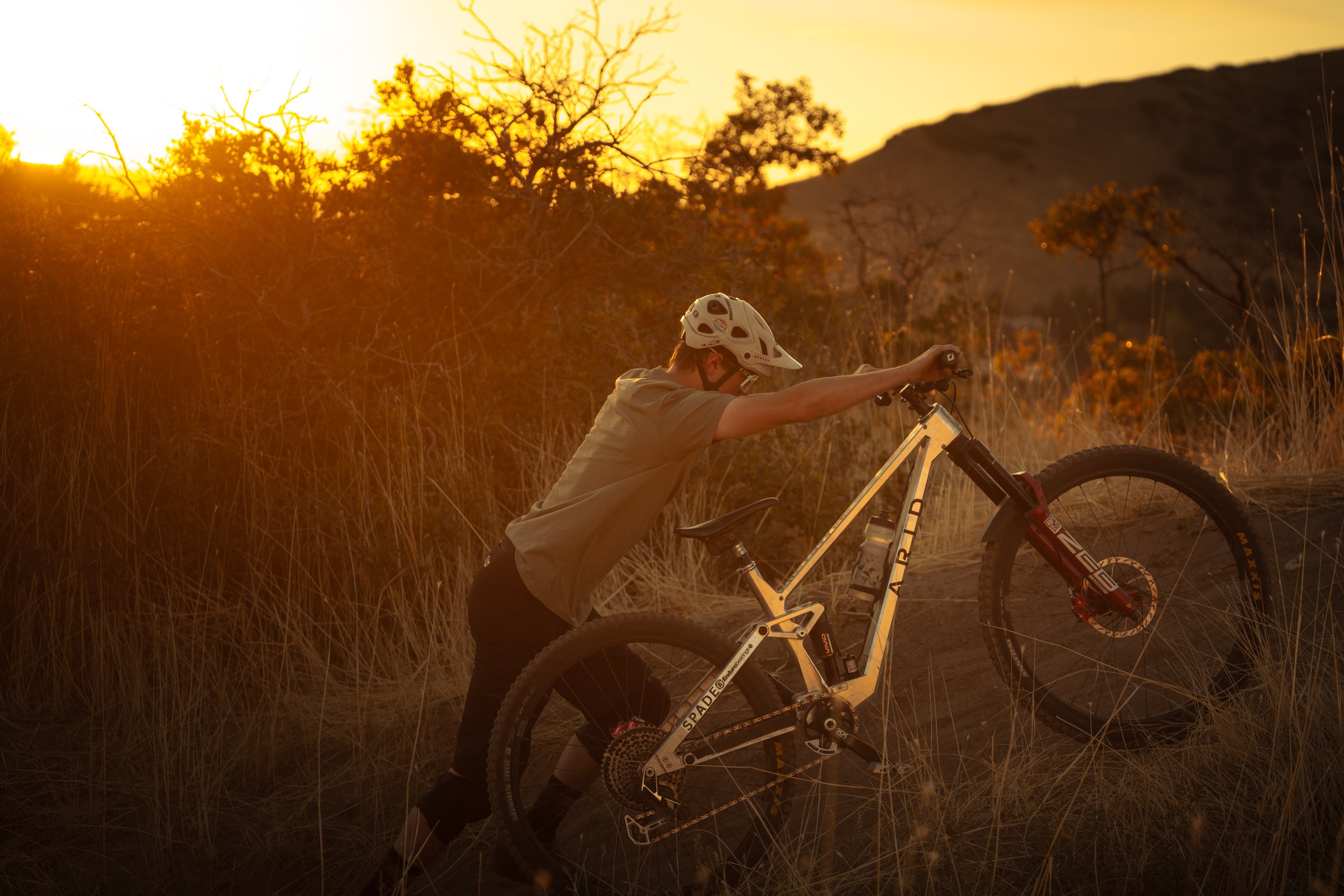 A person in a helmet pushing a mountain bike uphill during sunset in a grassy, hilly landscape. | Photography by Anders Henrikson Photo 