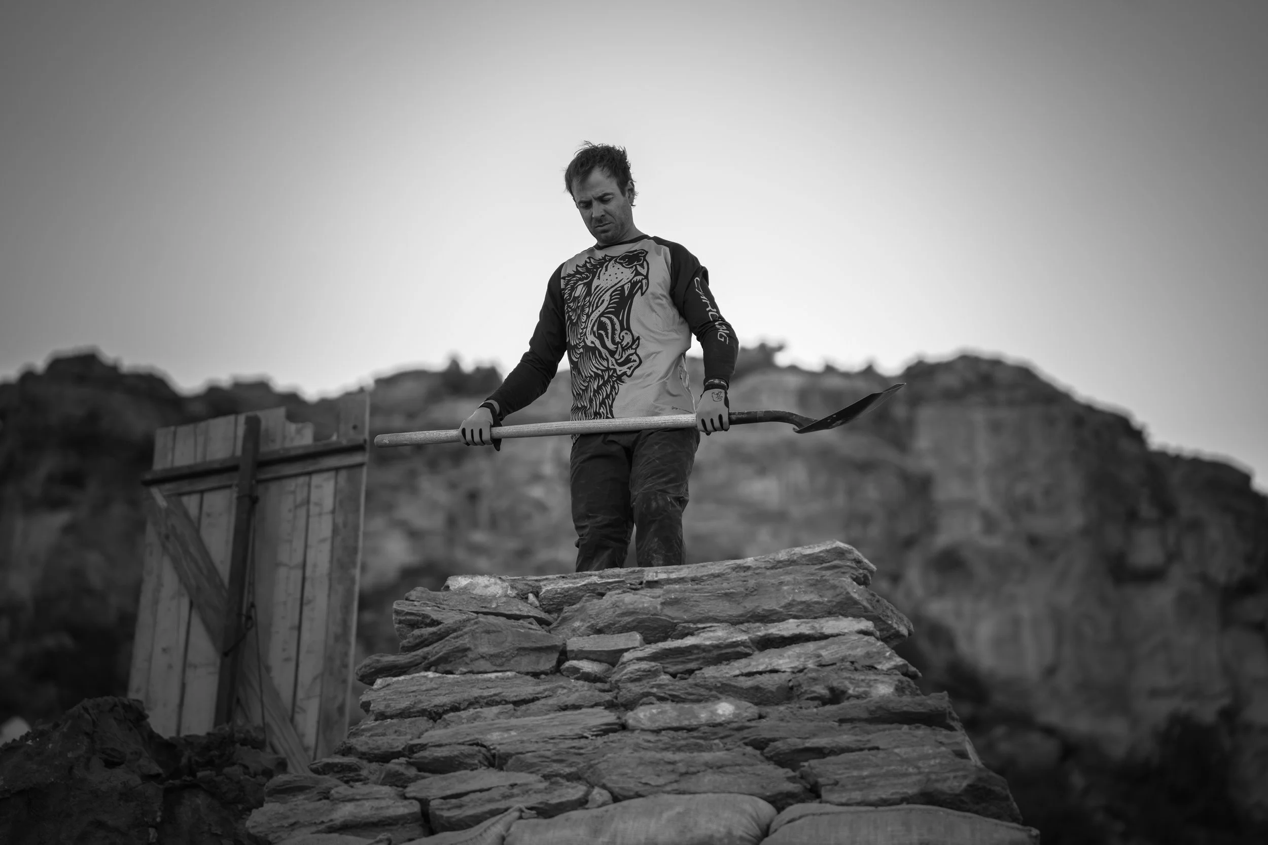 Person holding a shovel standing on a stone structure outdoors with rocky cliffs in the background.