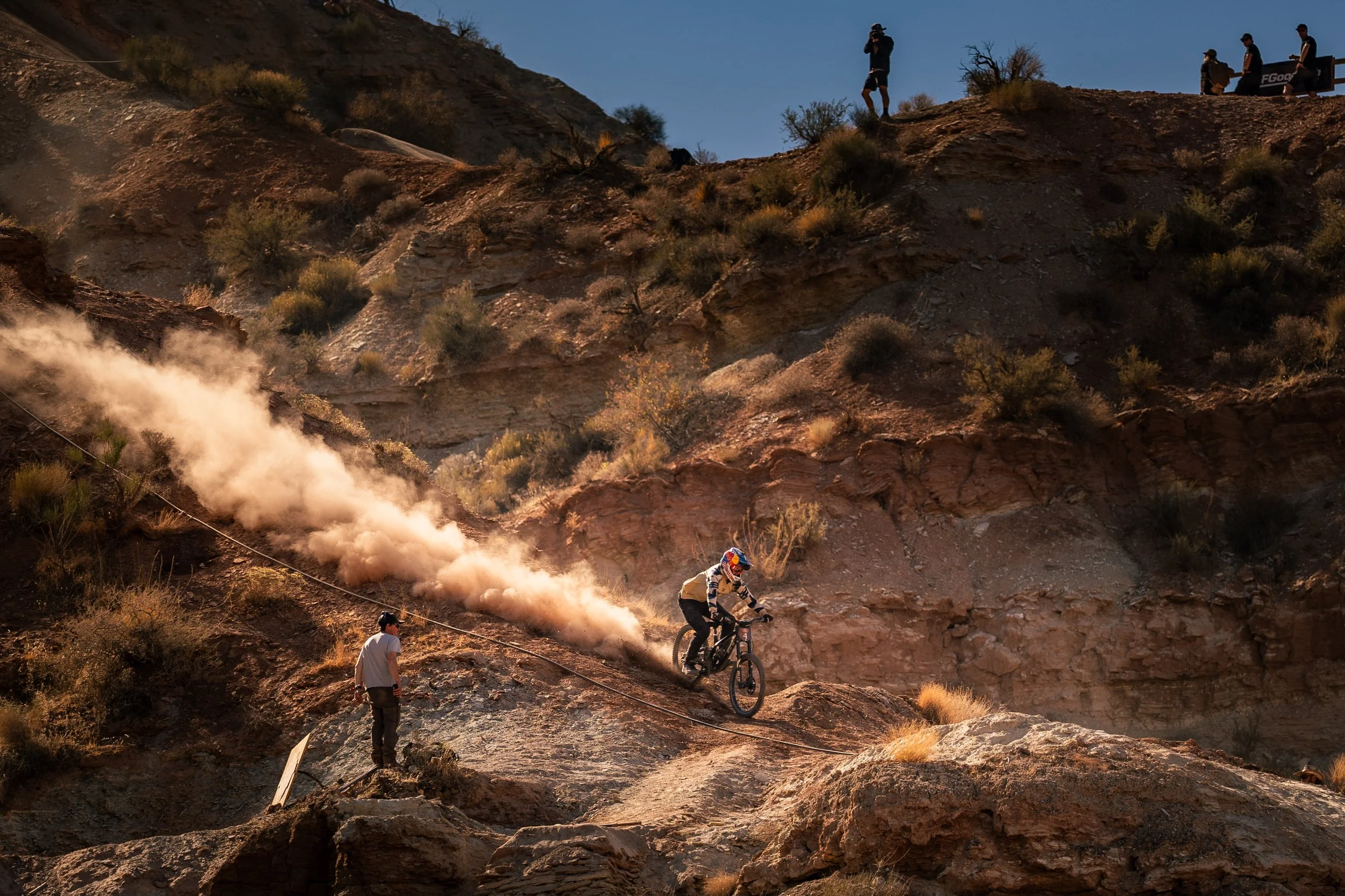 Mountain biker riding down a steep dirt trail in a rocky landscape, raising dust behind. Spectators are visible on top of the ridge.