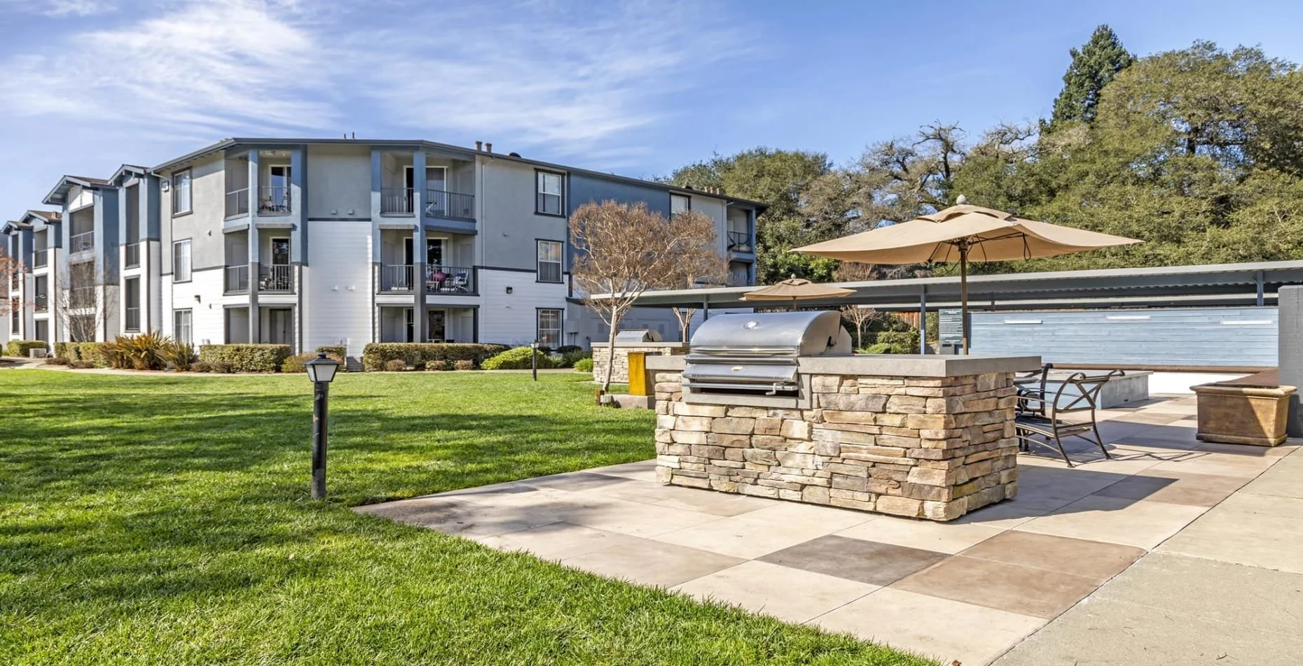 Outdoor shared patio area with a built-in grill, two umbrellas, seating, and a grassy lawn with apartment buildings in the background.