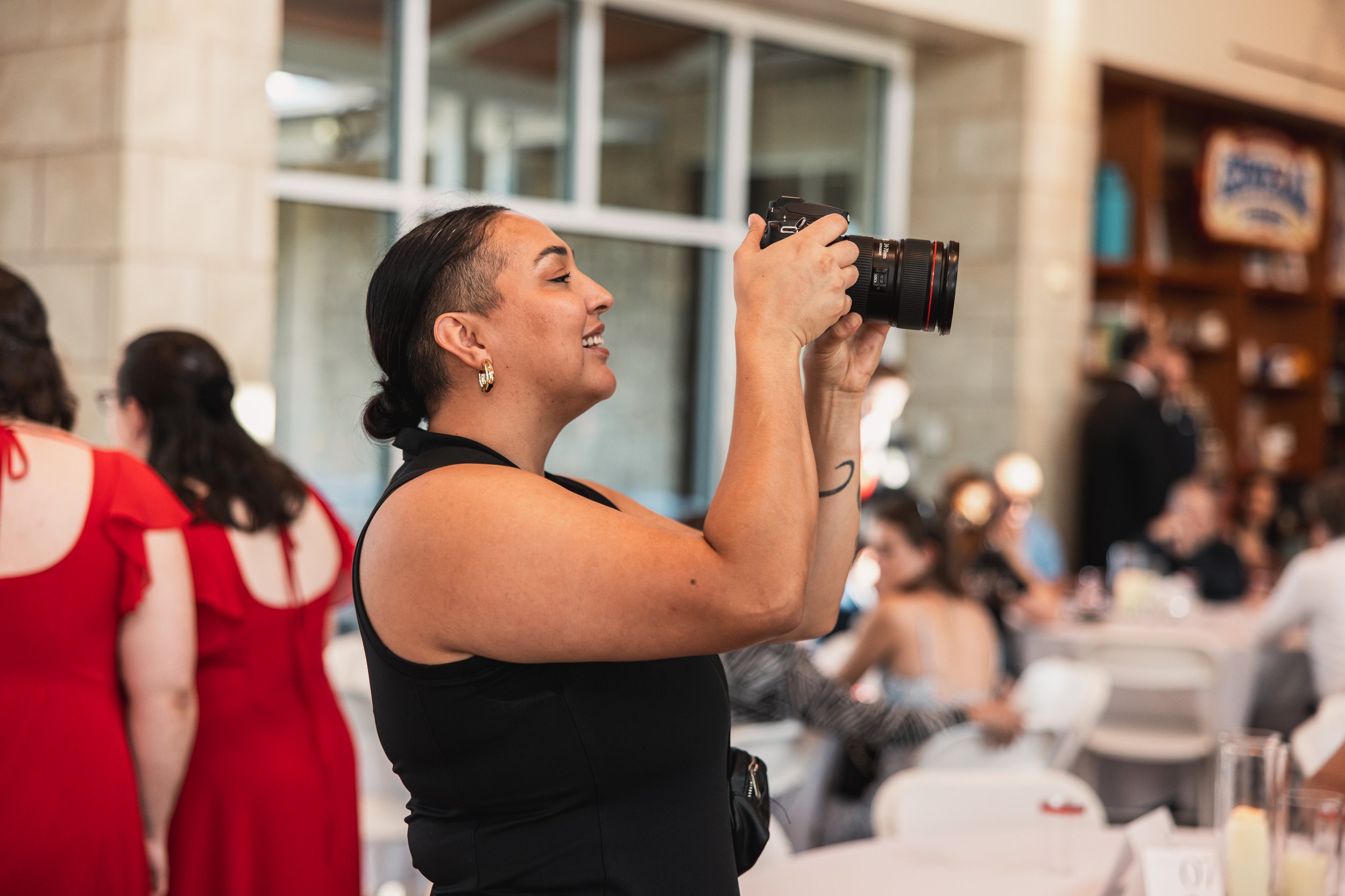 A woman with dark hair in a bun, wearing a black sleeveless top, taking a photograph with a camera at an indoor event with people seated at tables in the background.