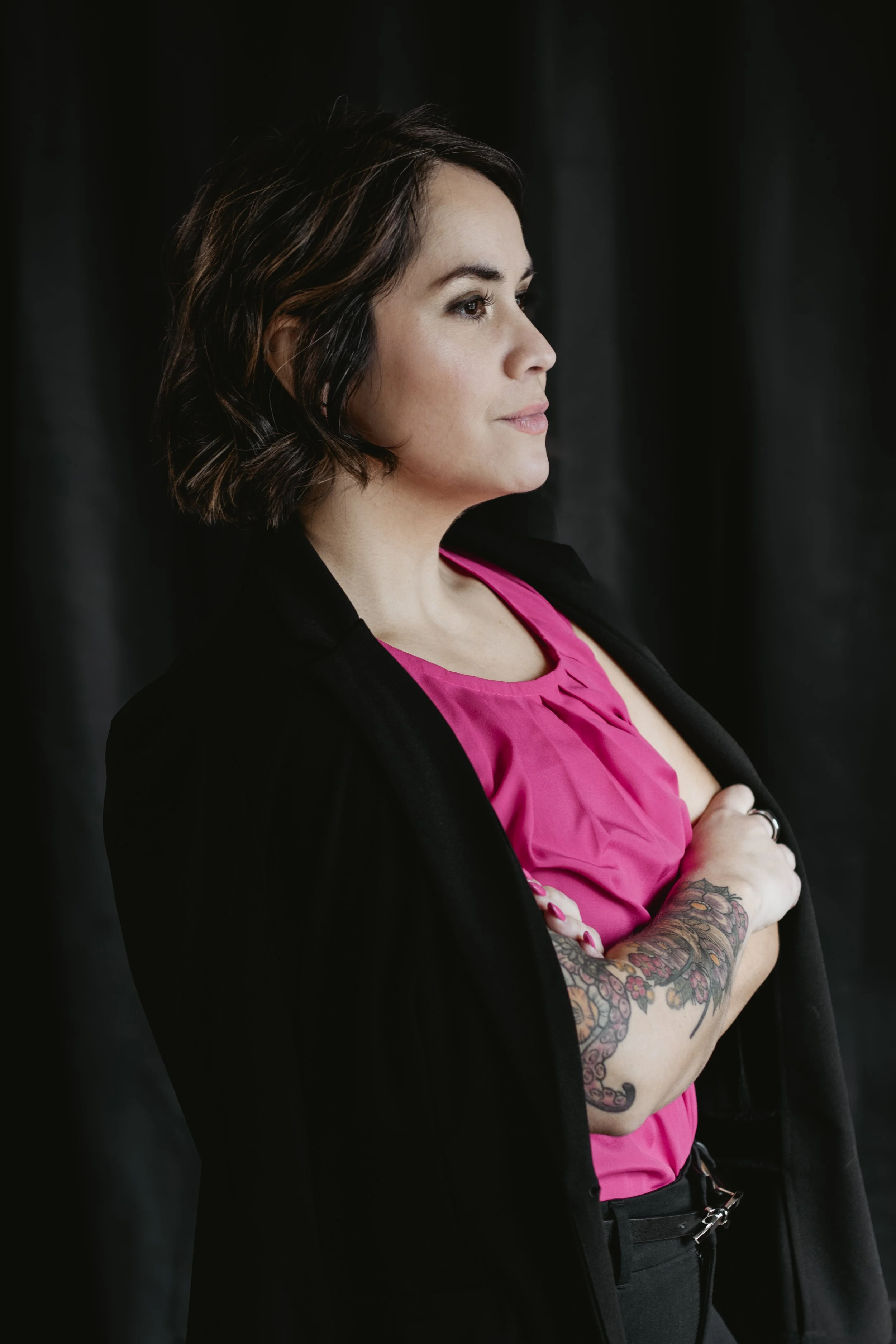 Smiling woman with short black hair, black top, with tattoos, standing against a white wall.