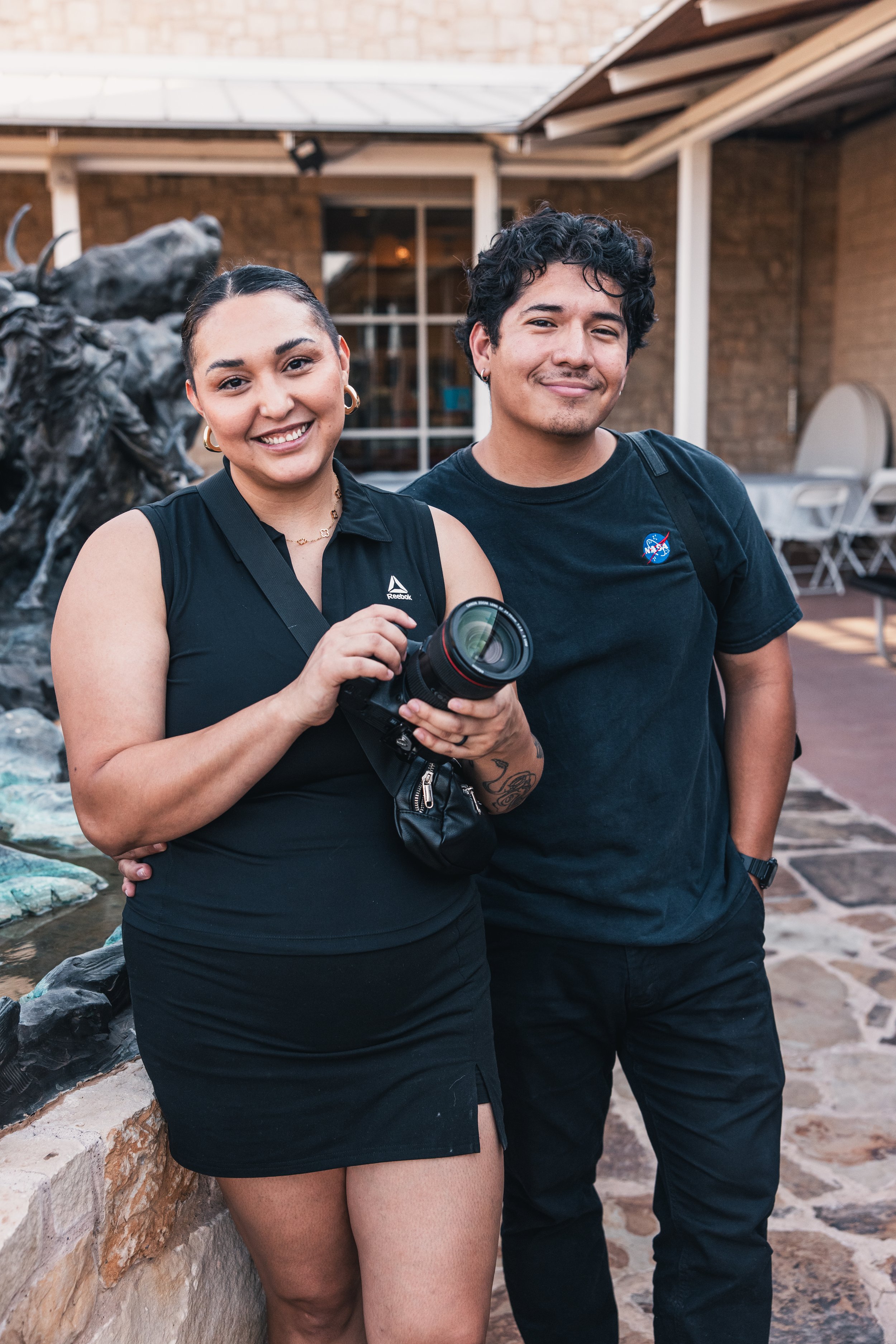 A woman and man standing outdoors at a social gathering, the woman holding a camera, both smiling at the camera.