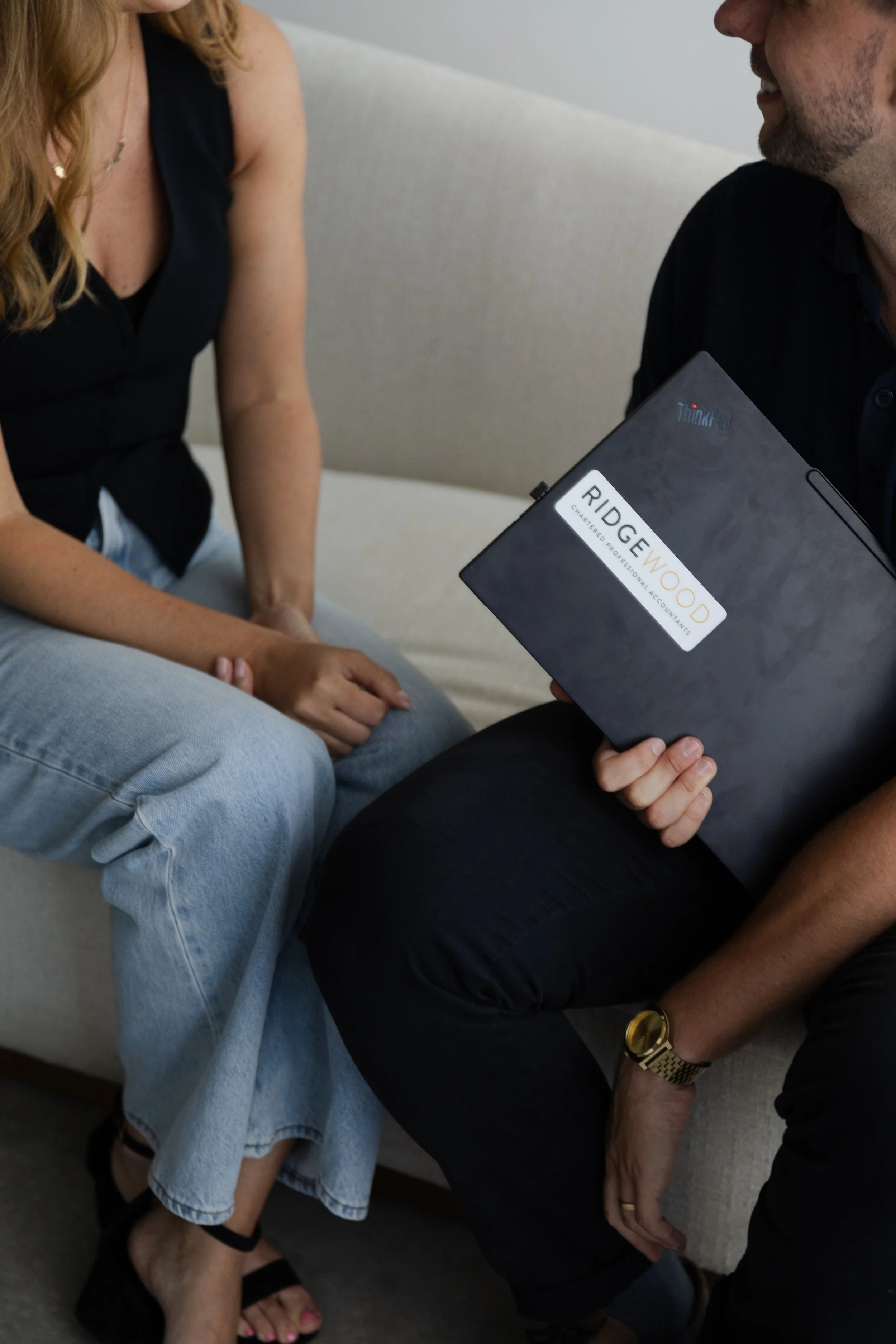 Two people sitting close together, one holding a laptop with a sticker that reads 'Ridgewood Chartered Professional Accountants.' The woman is wearing a sleeveless black top and light blue jeans, with sandals. The man is wearing a black shirt, dark pants, a gold watch, and a gold ring. They are engaged in conversation, sitting on a light-colored couch.
