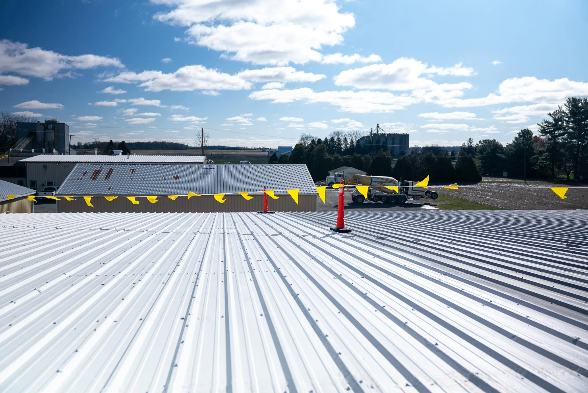 View of a rooftop with white metal roofing, yellow caution flags, and a construction trailer with a cement mixer attached. In the background, there are trees, farm buildings, and silos under a partly cloudy sky.