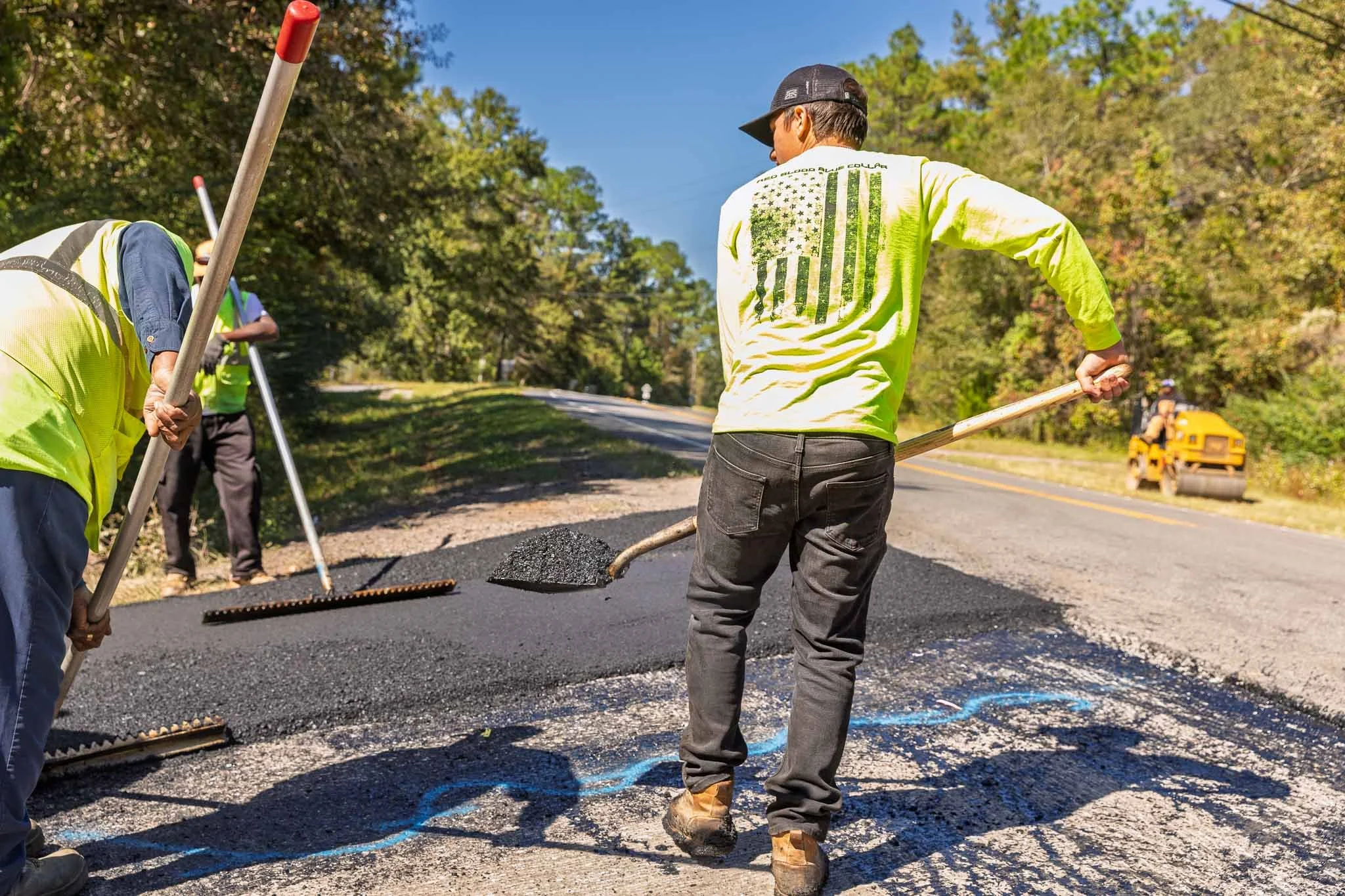 Workers paving a road with asphalt, using rakes and shovels, in a rural area surrounded by trees, with a yellow school bus in the background.