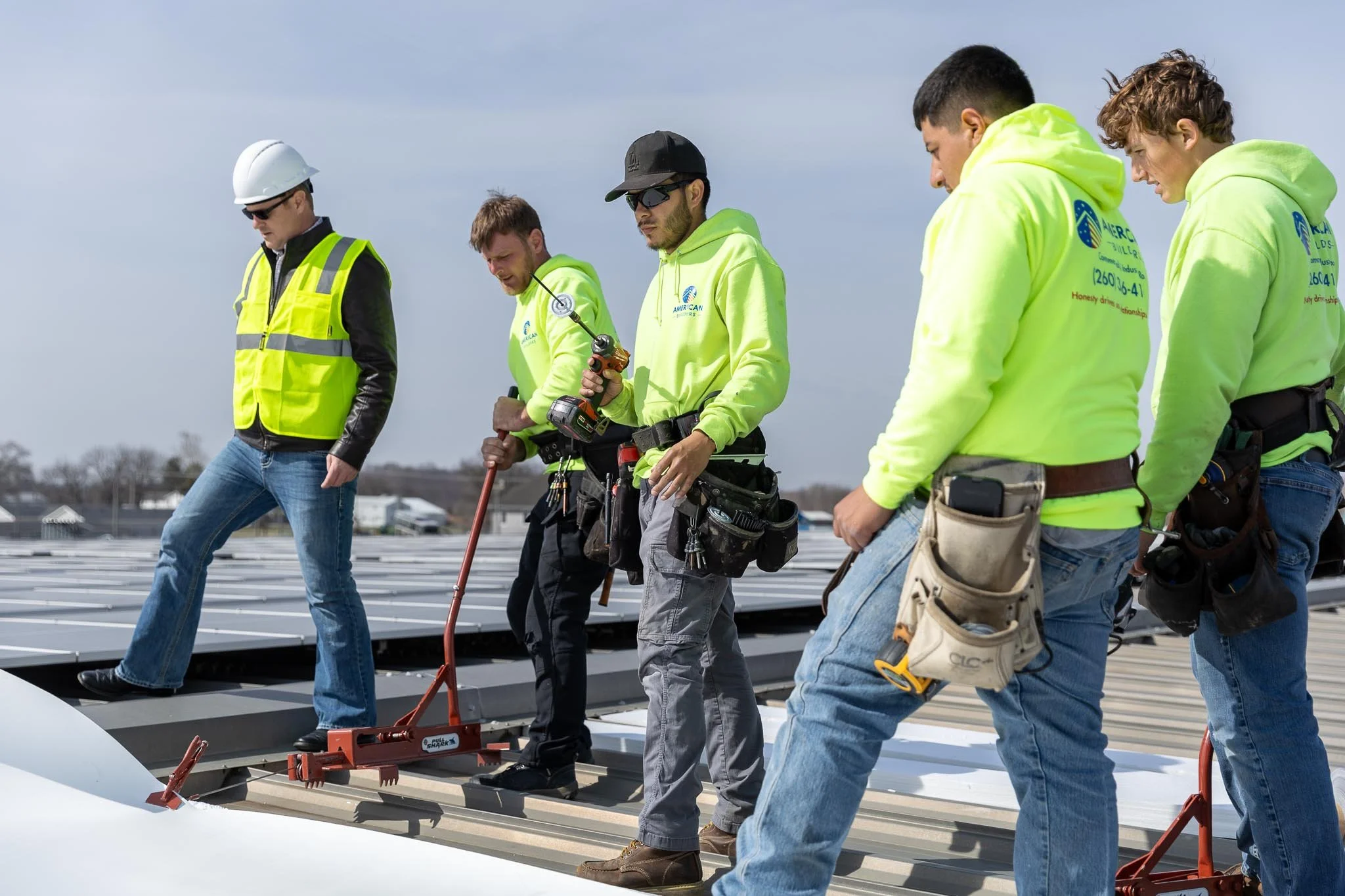 Group of construction workers wearing bright yellow safety hoodies and tools, working on roof installation under cloudy sky.