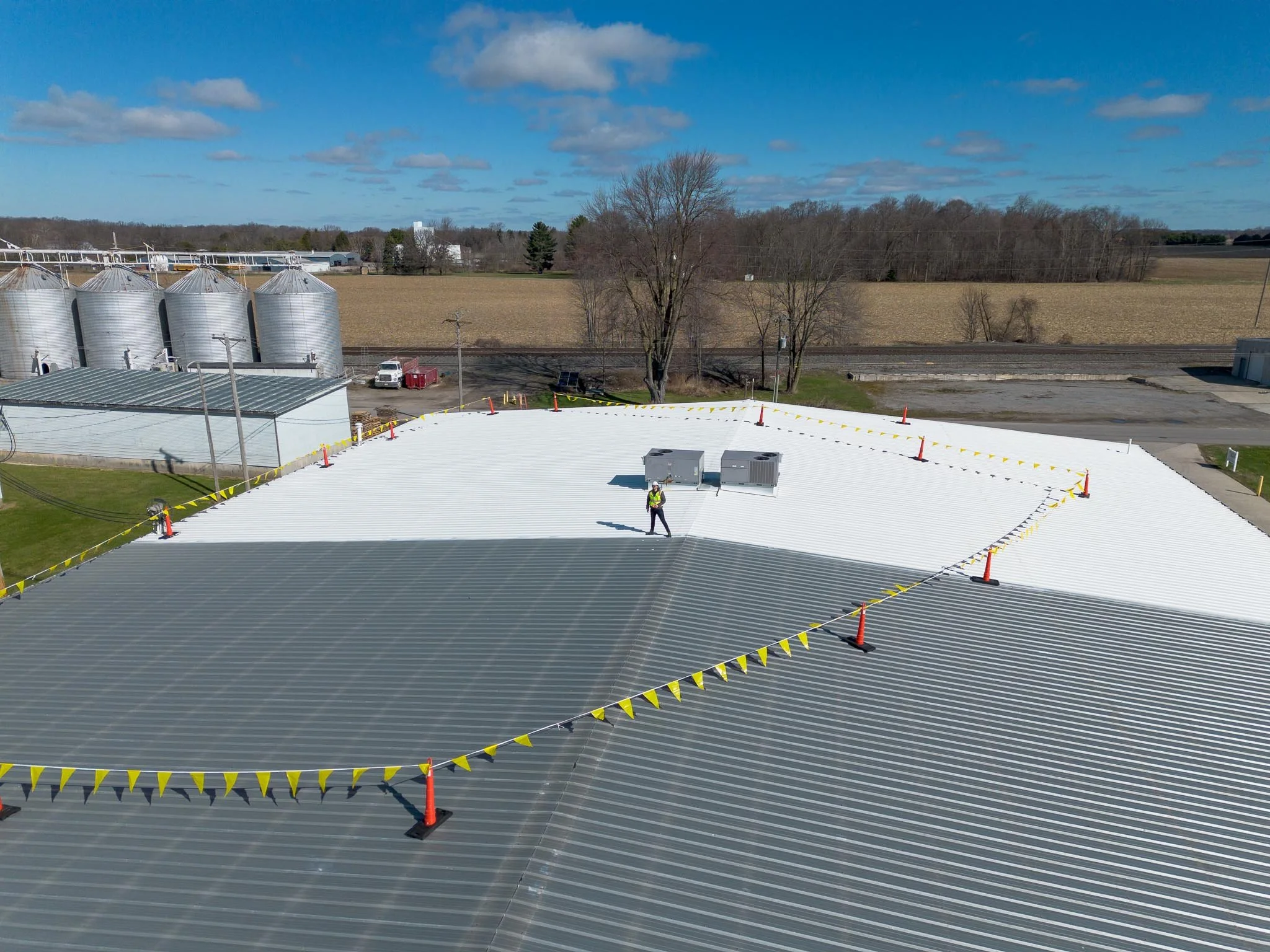 A person standing on a white rooftop with yellow caution tape, surrounded by orange traffic cones, with grain silos, trees, and open fields in the background under a partly cloudy sky.