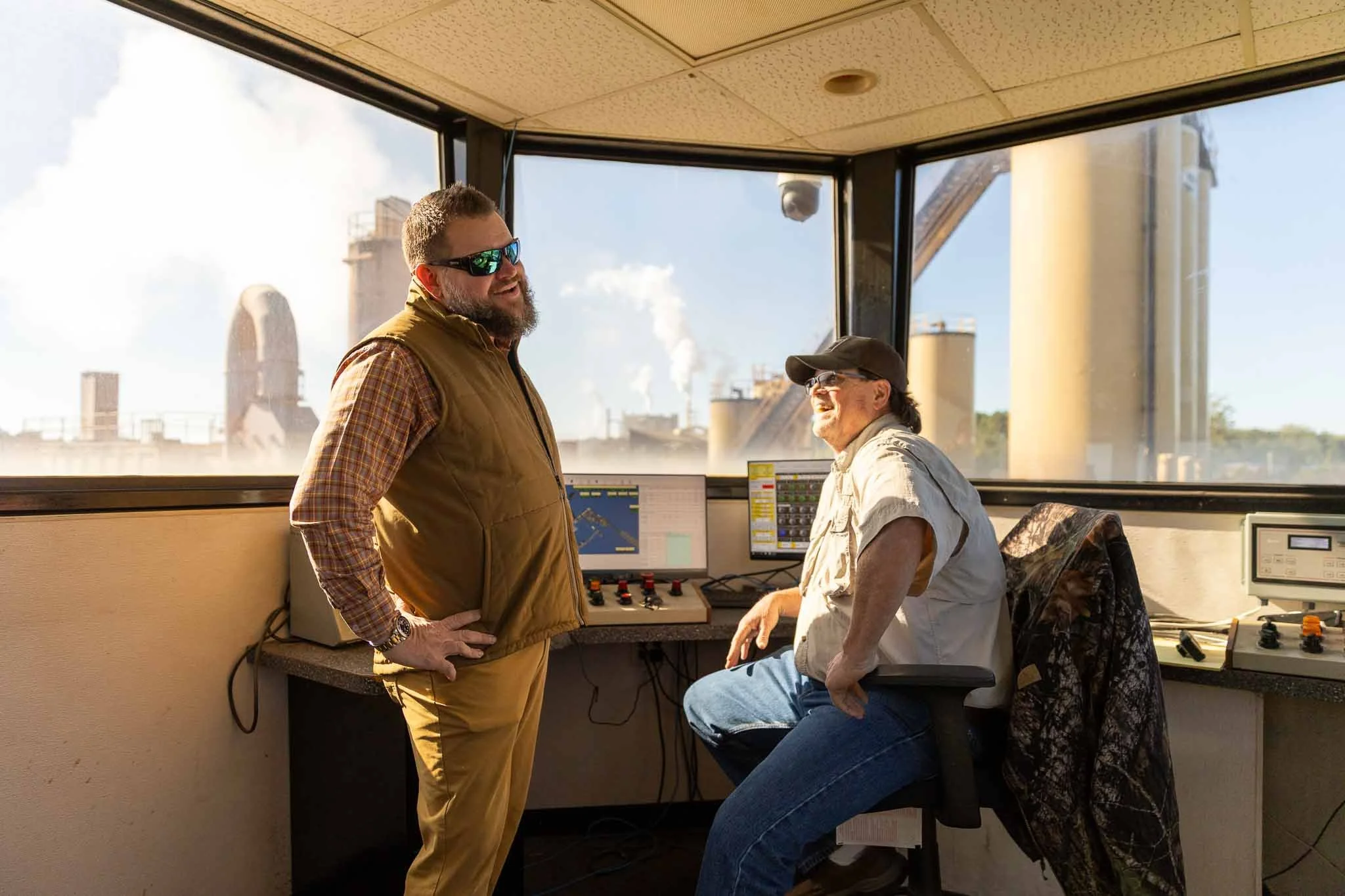 Two men are inside a room with large windows showing a cityscape with industrial buildings and smokestacks. One man is standing, wearing sunglasses and a brown vest, and the other is sitting at a control panel, wearing a cap and beige shirt. They app