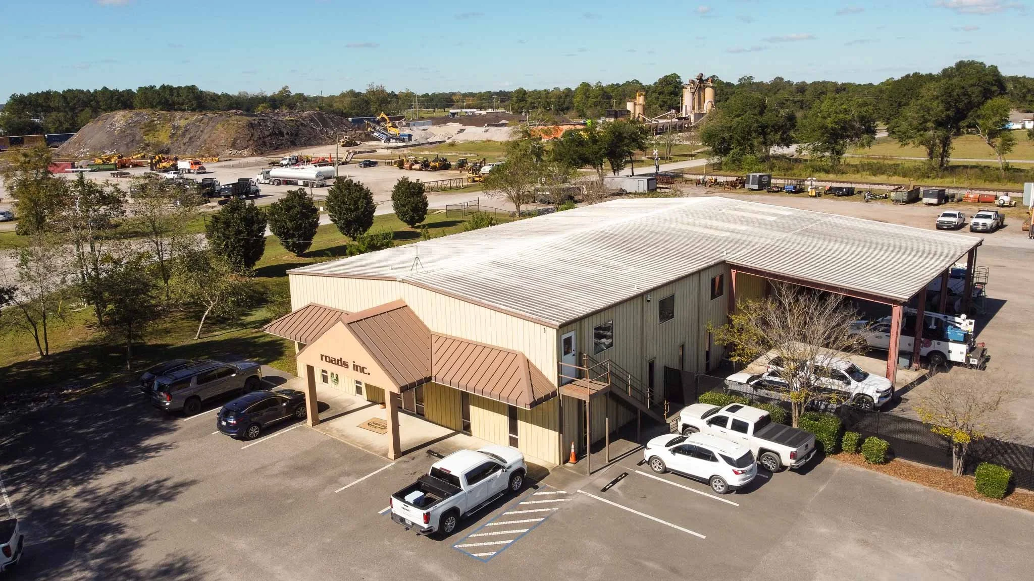 An aerial view of a parking lot with several cars, a building with a sign that says 'roads inc.', and a construction site with a large mound of dirt and construction equipment in the background. There are trees and grassy areas surrounding the parkin