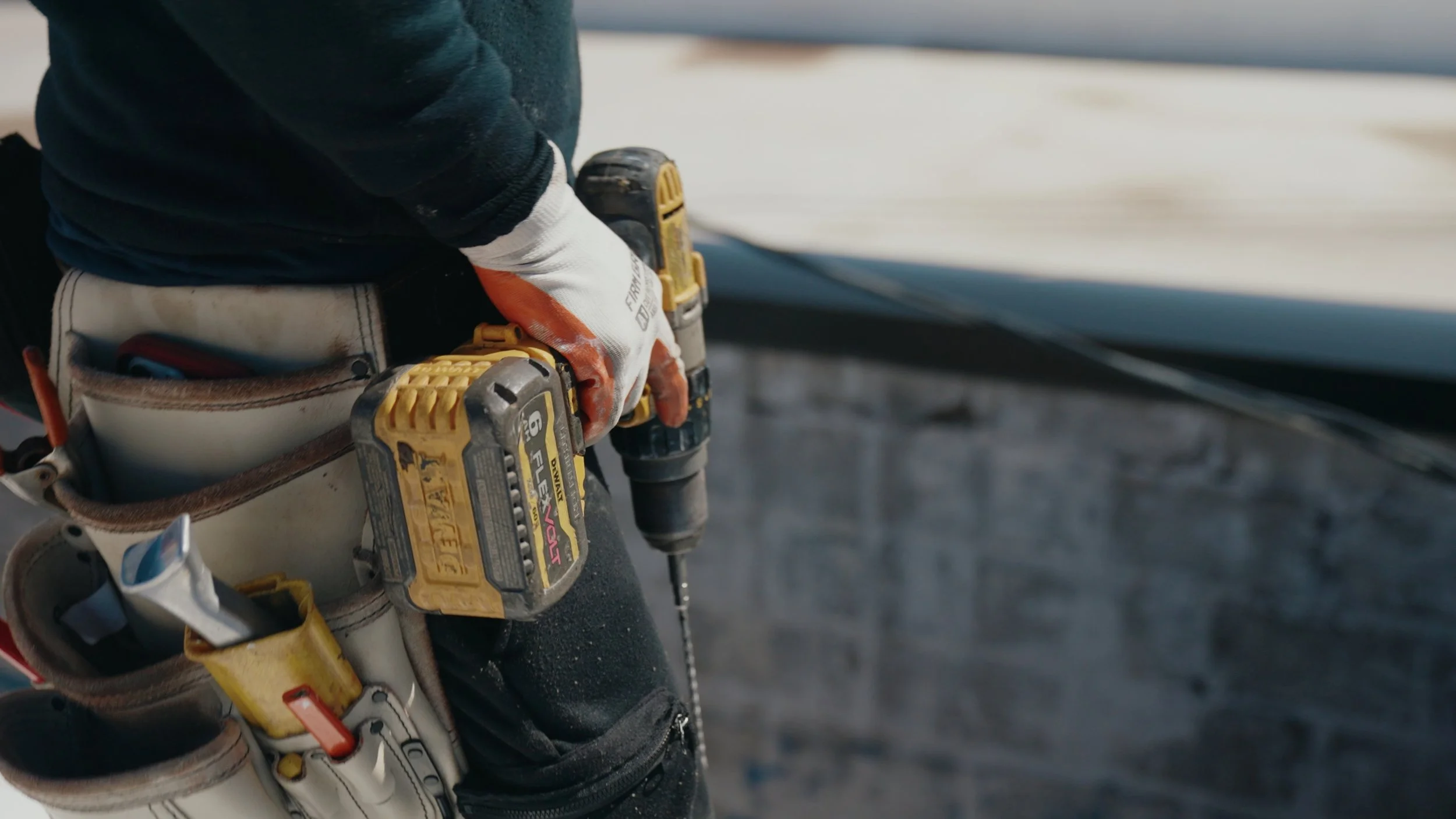 Close-up of a worker's gloved hand operating a yellow cordless drill, with additional tools and a utility belt around their waist, on a construction site.