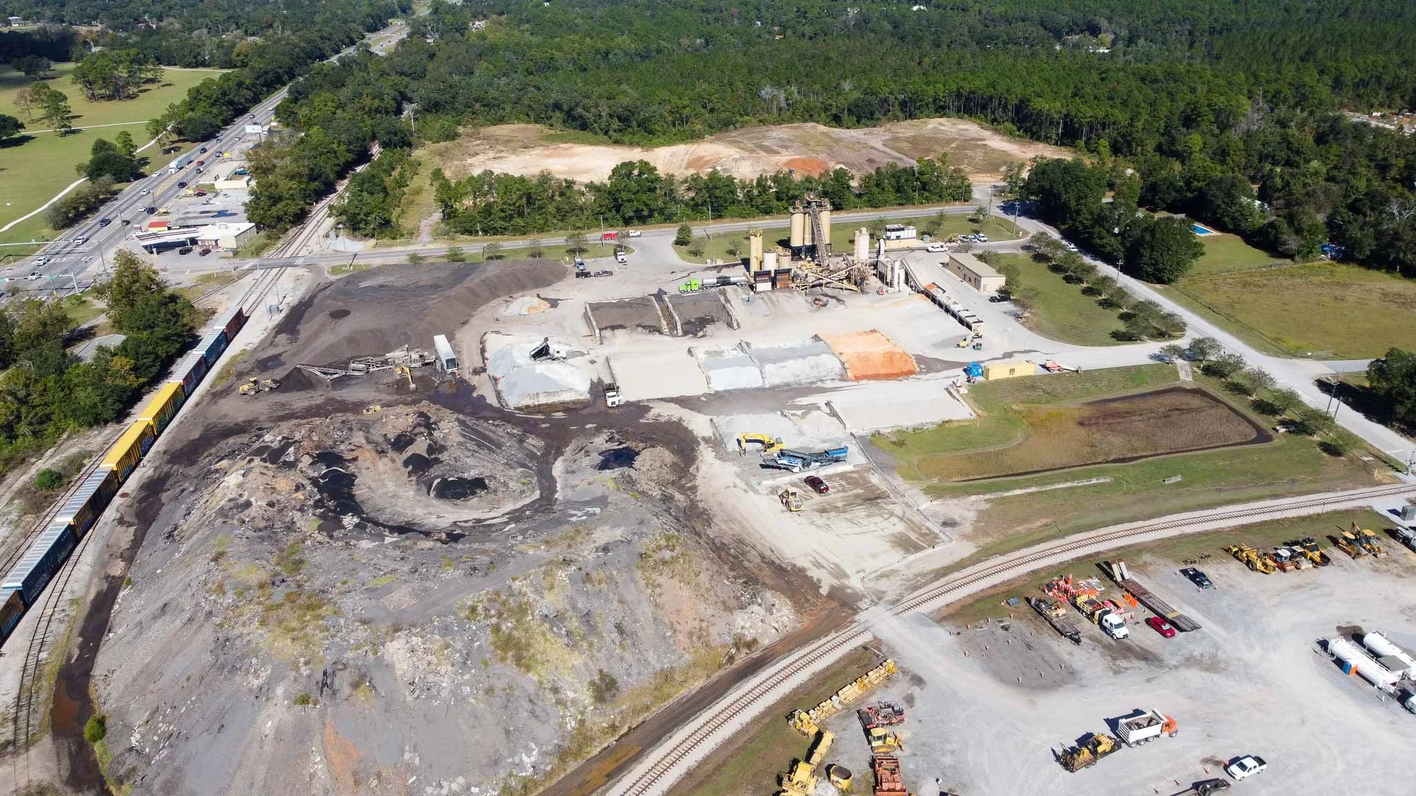 An aerial view of a construction site next to a railroad track, with construction equipment, vehicles, and cleared land, surrounded by green trees and a small residential area in the distance.