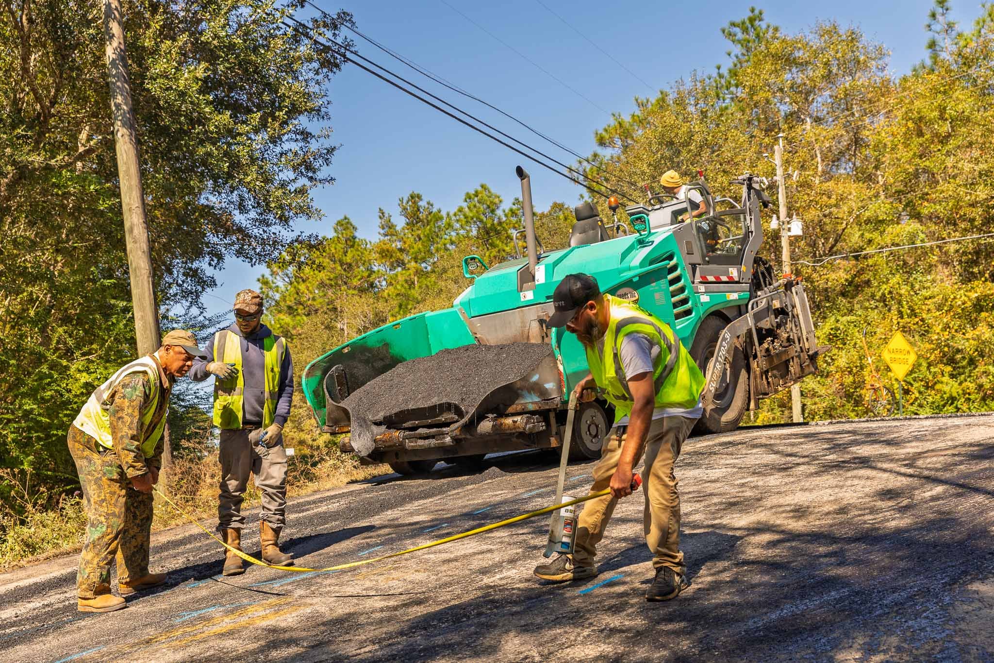Workers in safety vests and hats repair a section of a paved road with a paving machine under a blue sky, surrounded by trees.
