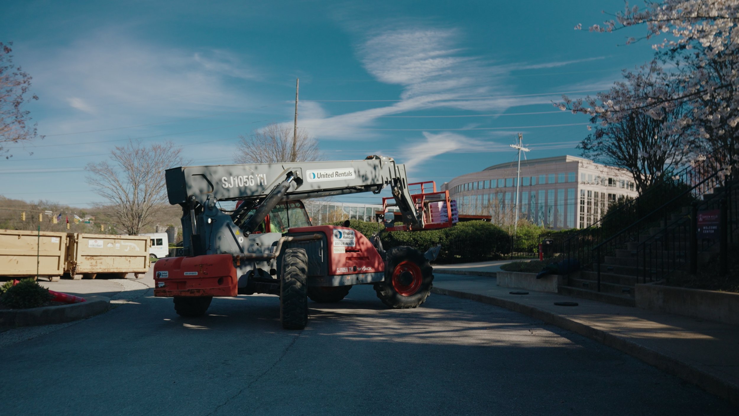 Construction lift truck parked outdoors beneath a partly cloudy blue sky, with cherry blossom trees and a modern glass building in the background.