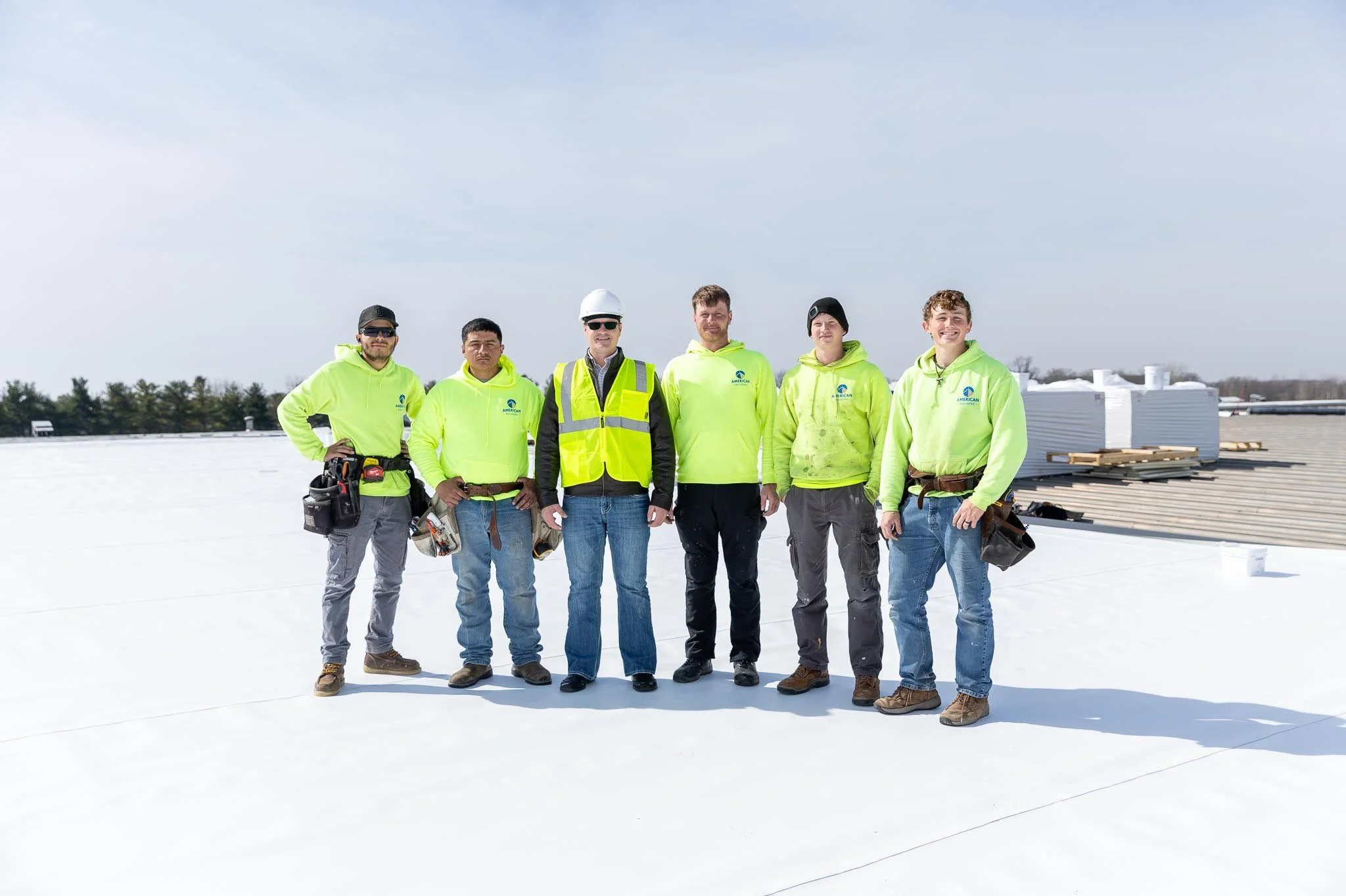 Group of six construction workers standing on a rooftop, posing for a photo. They are wearing high-visibility jackets, some with tools attached, and are dressed for cold weather with hats and gloves. The background shows a clear sky and some roofing 