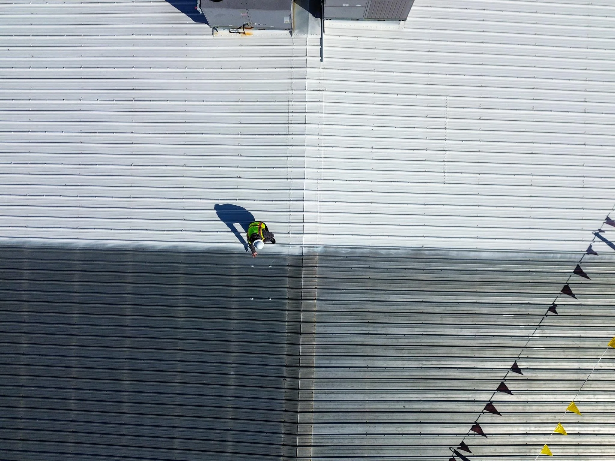 Bird's eye view of a person in a high-visibility vest and white hard hat walking on the roof of a large industrial building made of metal panels, with shadows cast on the roof surface.