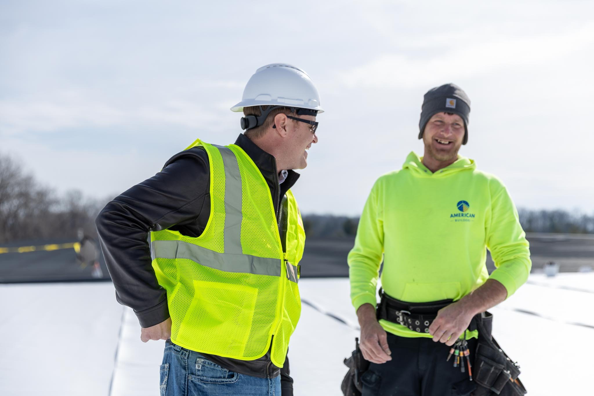 Two men in safety gear standing outdoors in a snowy area, smiling and talking.