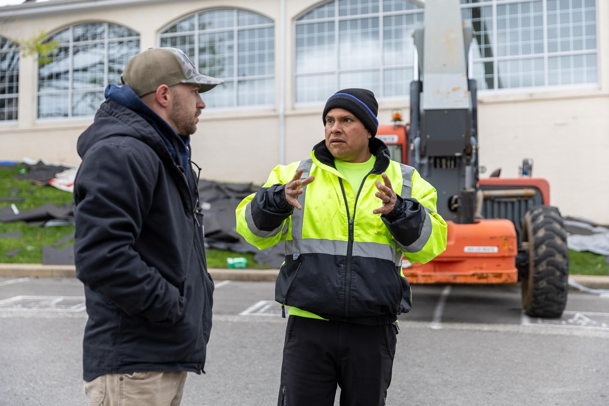 Two men engaged in conversation outdoors, one in a black jacket and beige cap, the other in a neon yellow safety jacket and black beanie, with construction equipment and a building with large windows in the background.