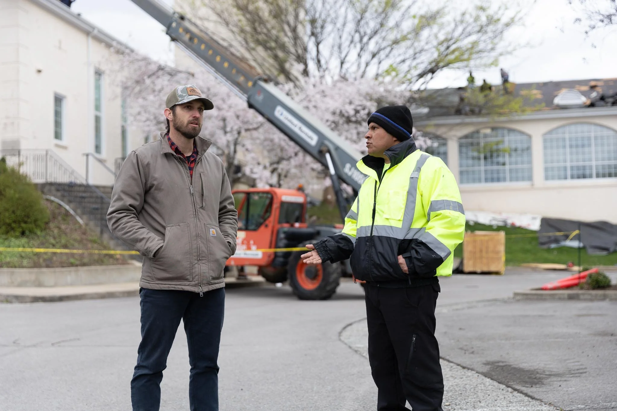 Two men are standing and talking outdoors near a construction site. One man is wearing a gray jacket and a baseball cap, while the other man is wearing a black beanie and a bright yellow safety jacket. There is construction equipment in the backgroun
