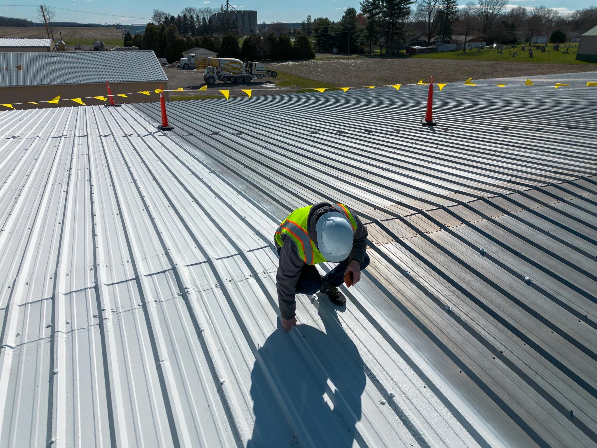 A worker wearing a safety helmet and reflective vest bending down on a metal roof, with orange and yellow safety cones and tape nearby. There are trees, fields, and a few buildings in the background, under a clear sky.