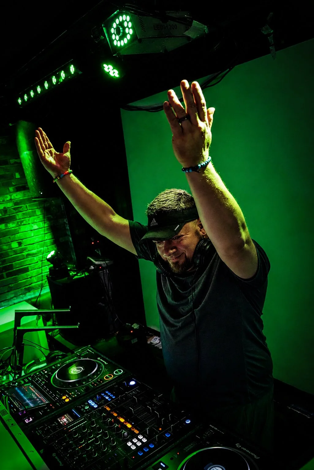 Trey Triplicate DJ'ing wearing a black visor and headphones around his neck, is smiling and raising his arms with hands open. He is standing behind a DJ booth with equipment, in a room illuminated with green lighting and a brick wall background.