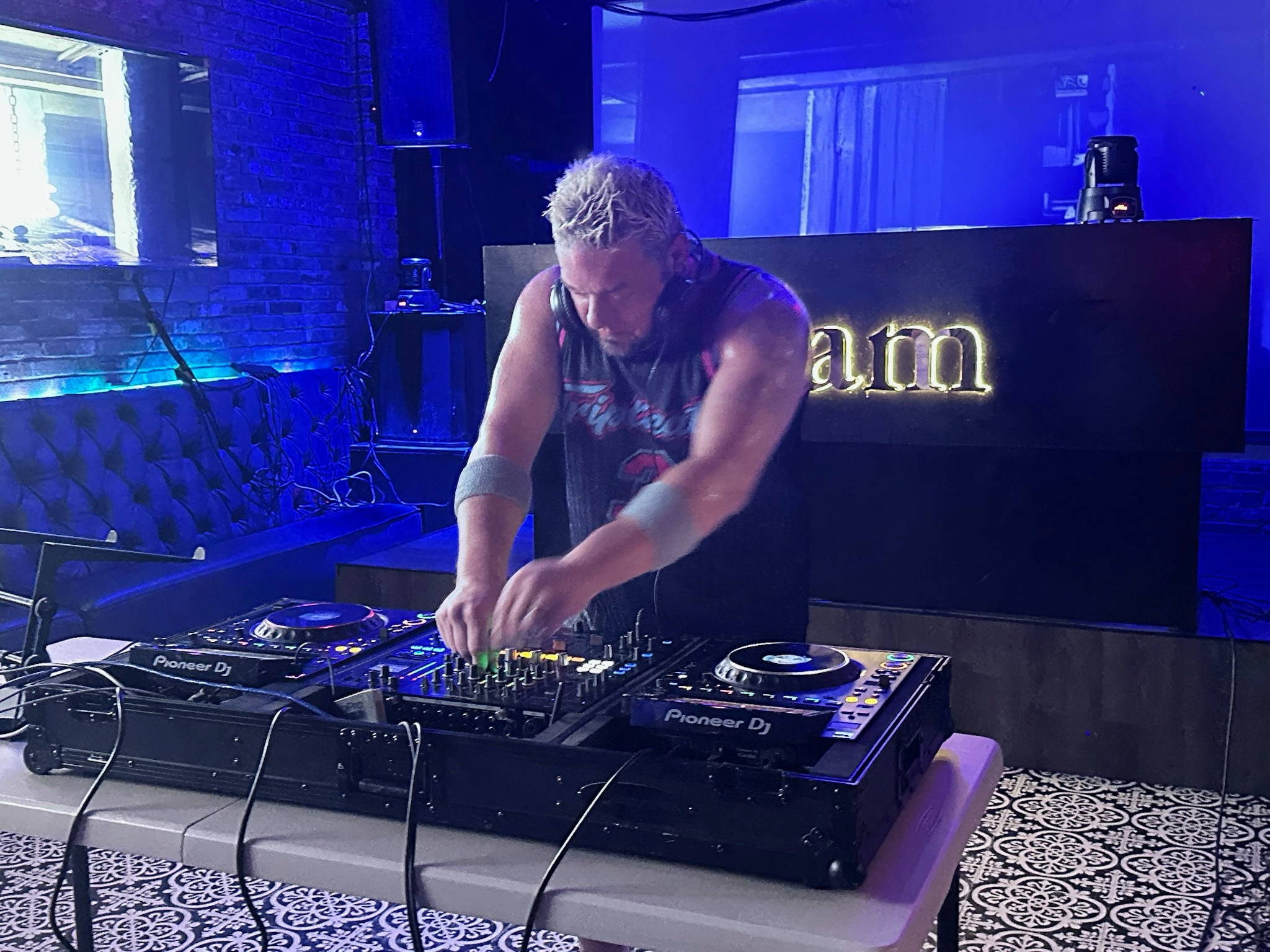Trey Triplicate, a DJ and producer from atlanta, georgia is wearing headphones is performing behind DJ equipment on a stage with blue lighting. The background features a black brick wall, a large screen, with letters behind him that say GLAM
