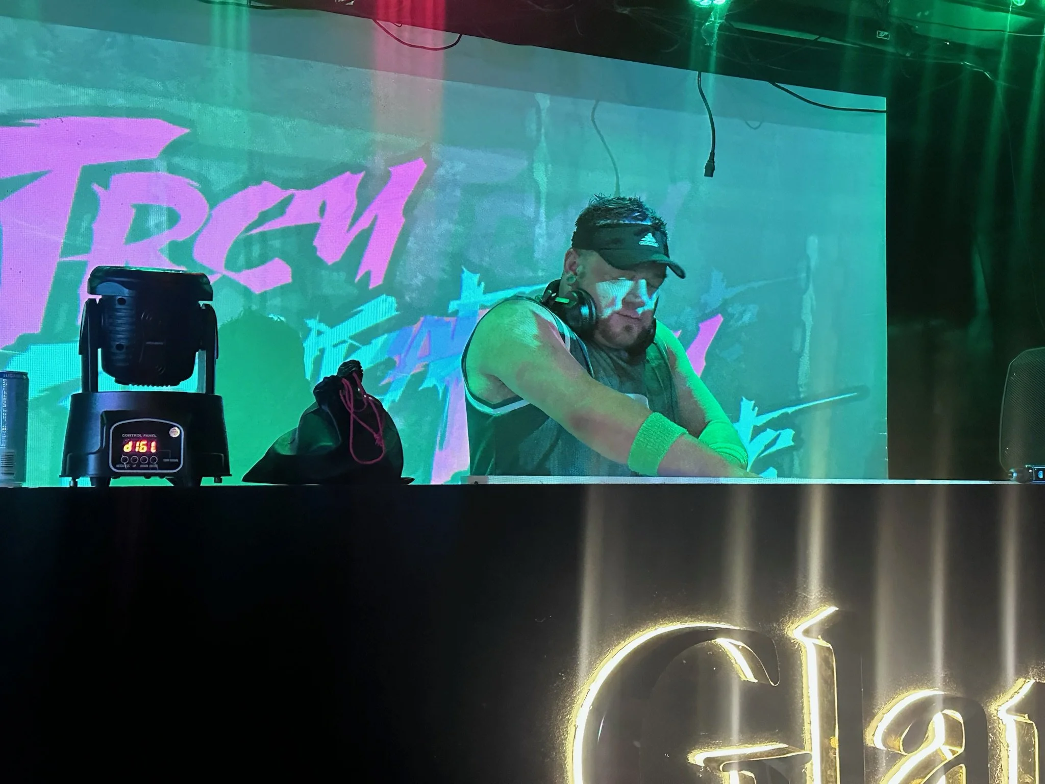 International Superstar DJ Trey Triplicate with headphones and sunglasses is DJing at a colorful, vibrant club or event with bright lights and a large screen behind him displaying the word "TREY TRIPLICATE" in stylized pink and cyan text.