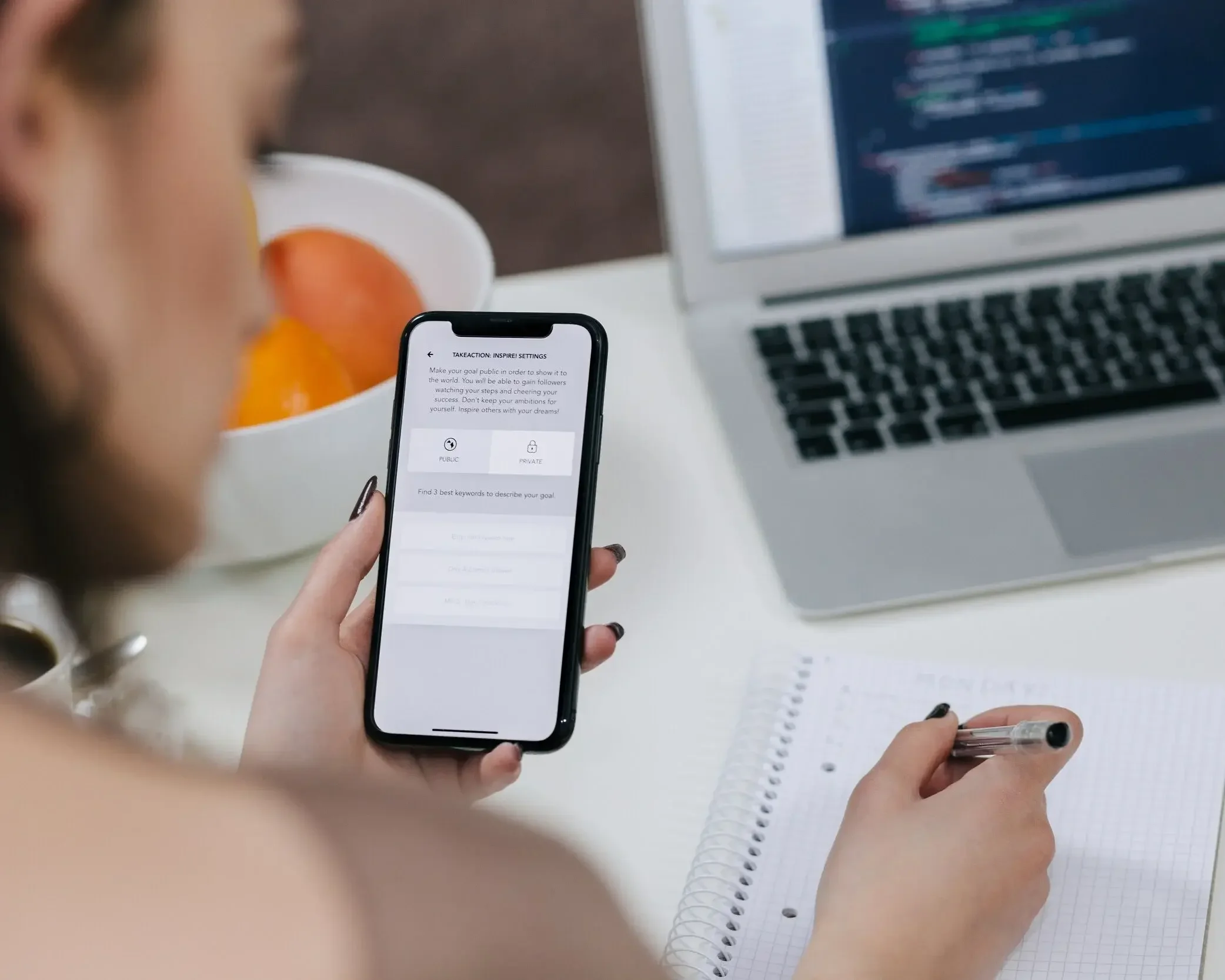 A person holding a smartphone displaying a goal setting app, with a computer, a notebook, and a bowl of oranges on the desk.