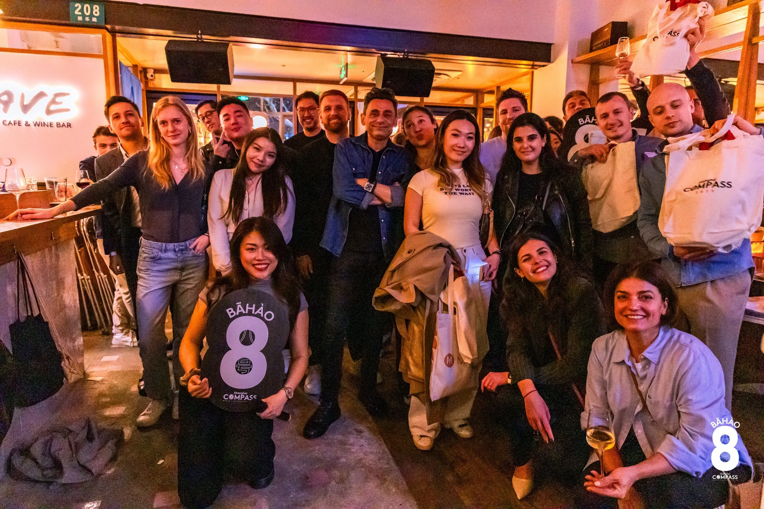 A group of people posing for a photo at a bar or restaurant, with some holding drinks and one person holding a sign with the number 8. The setting appears to be lively and social, with warm lighting and a visible sign that reads 'LOVE' in the backgro