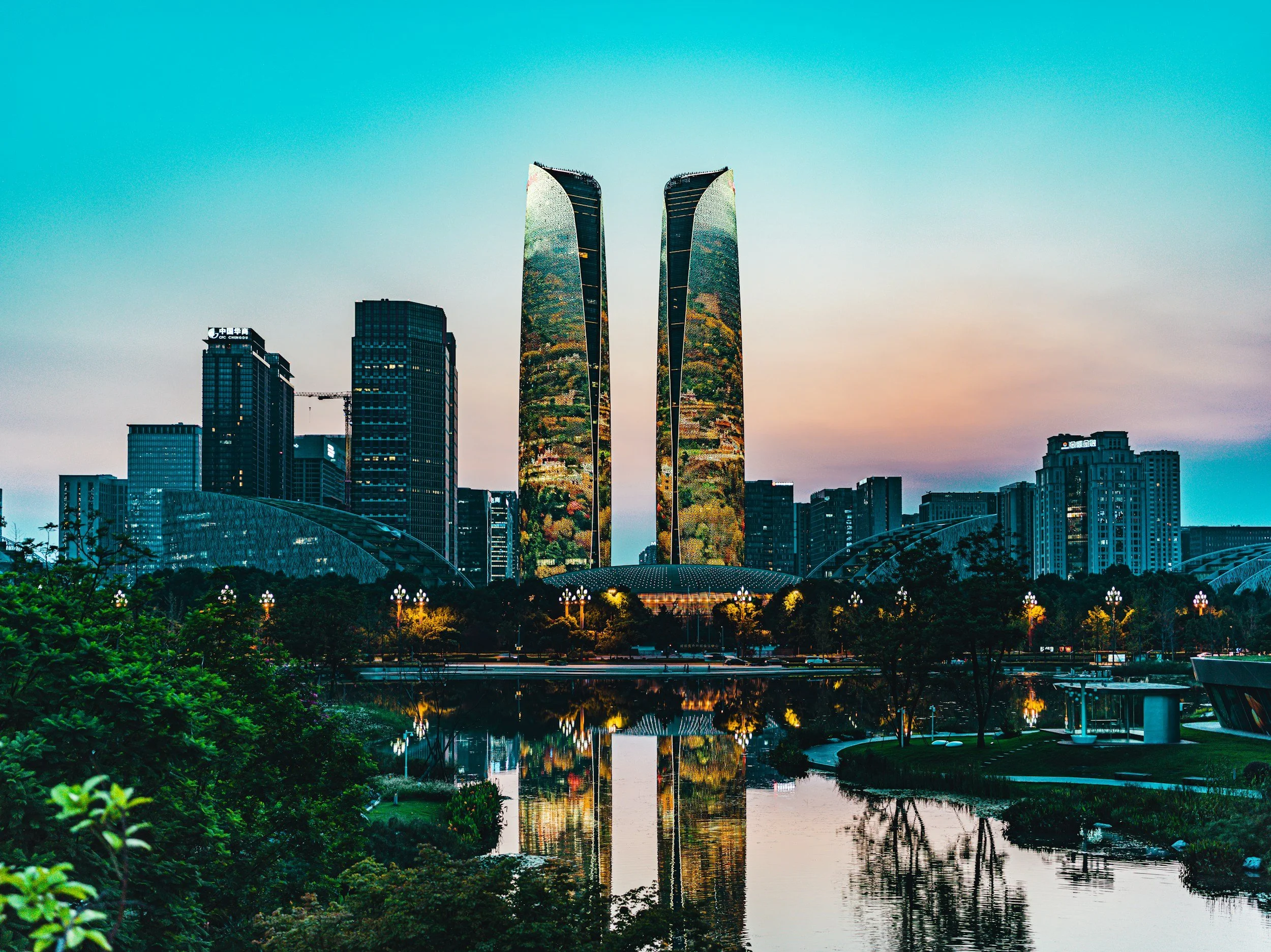 City skyline at dusk featuring modern skyscrapers, including two tall, curved glass towers, reflected in a river with greenery in the foreground.
